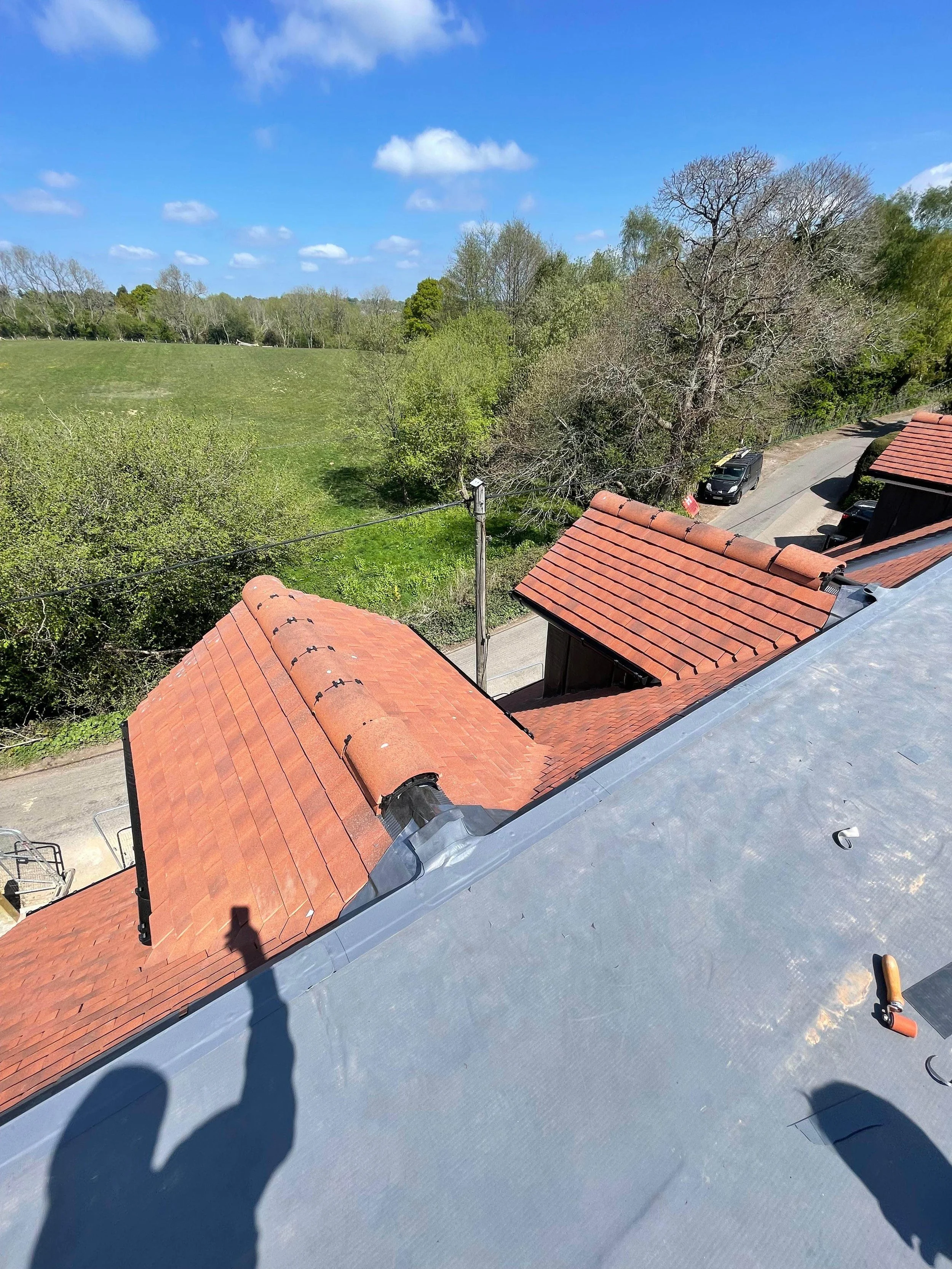 View from a rooftop showing red tile roofs, a street with parked cars, trees, green field, and a partly cloudy sky.