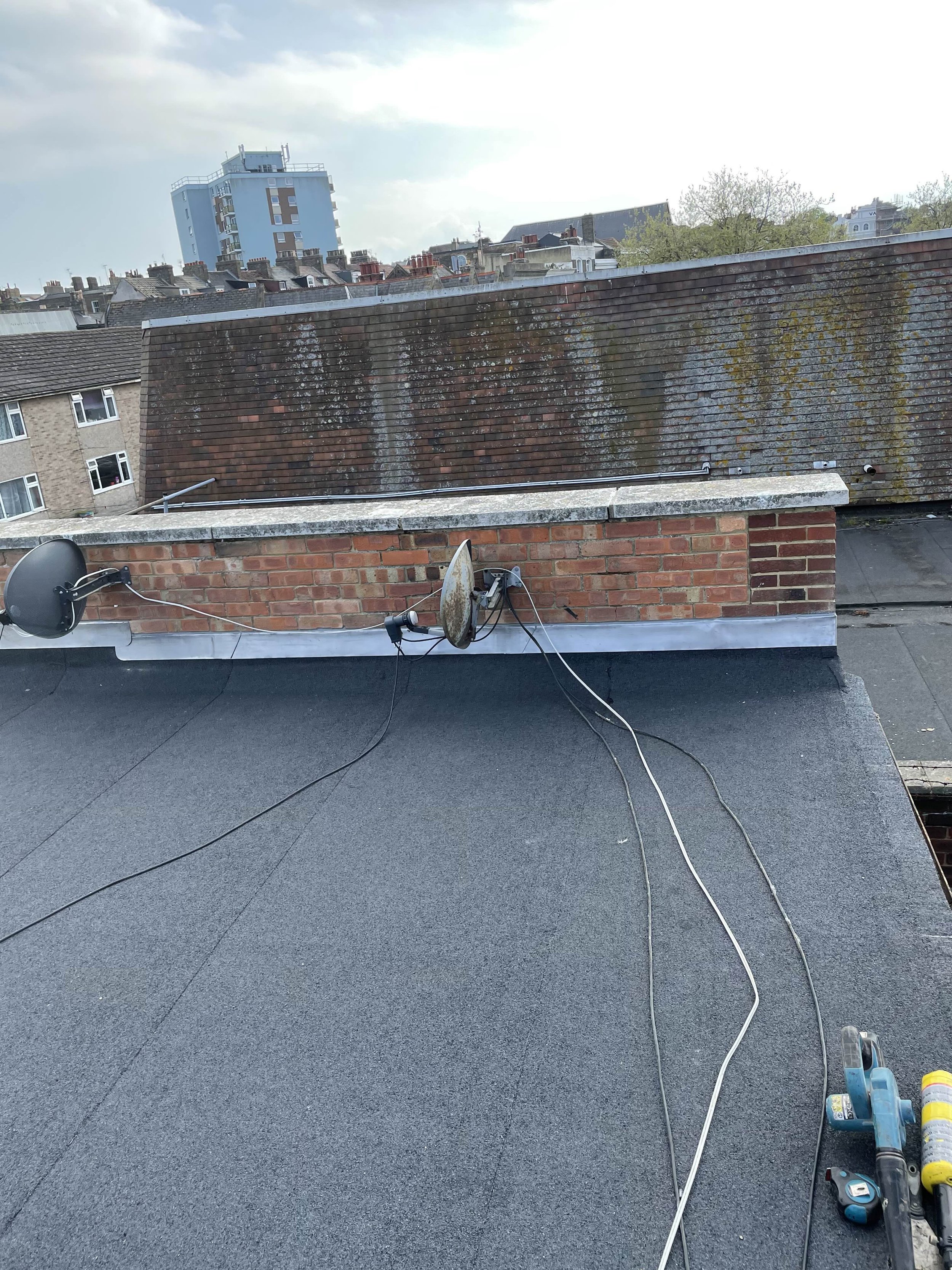 View of rooftops showing a chimney with two satellite dishes and wires, in an urban area with other buildings in the background.