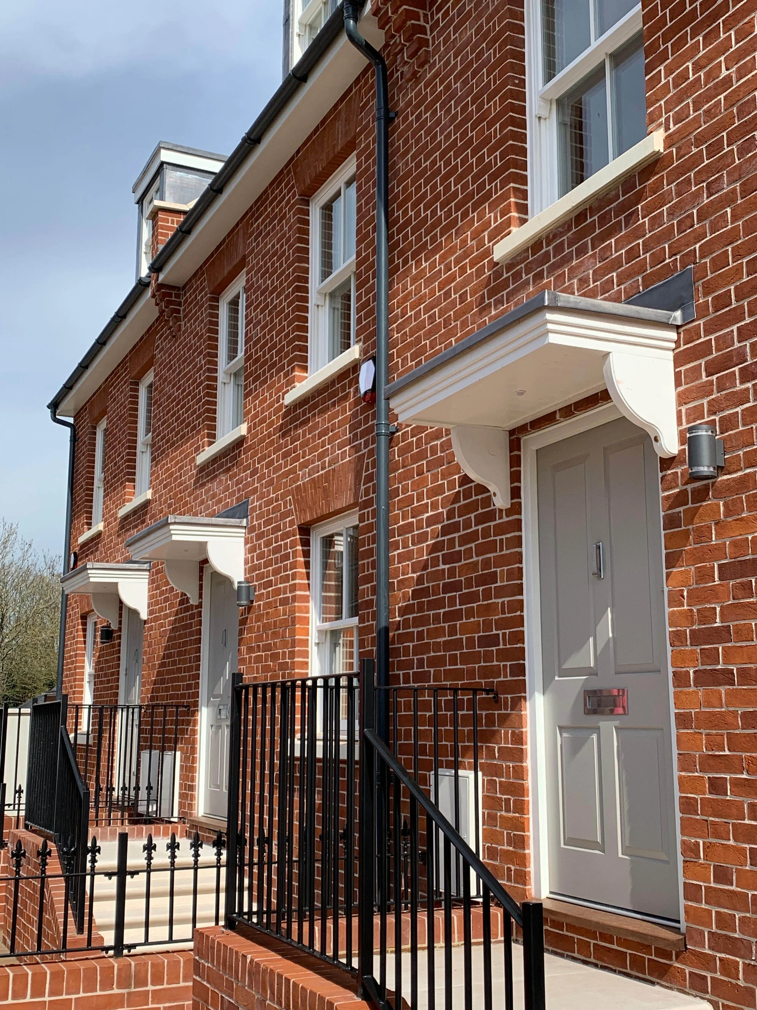 Exterior view of a red brick townhouse with white doors and small porches, black metal railings, black downspouts, and three windows with white trim, under a sunny sky.
