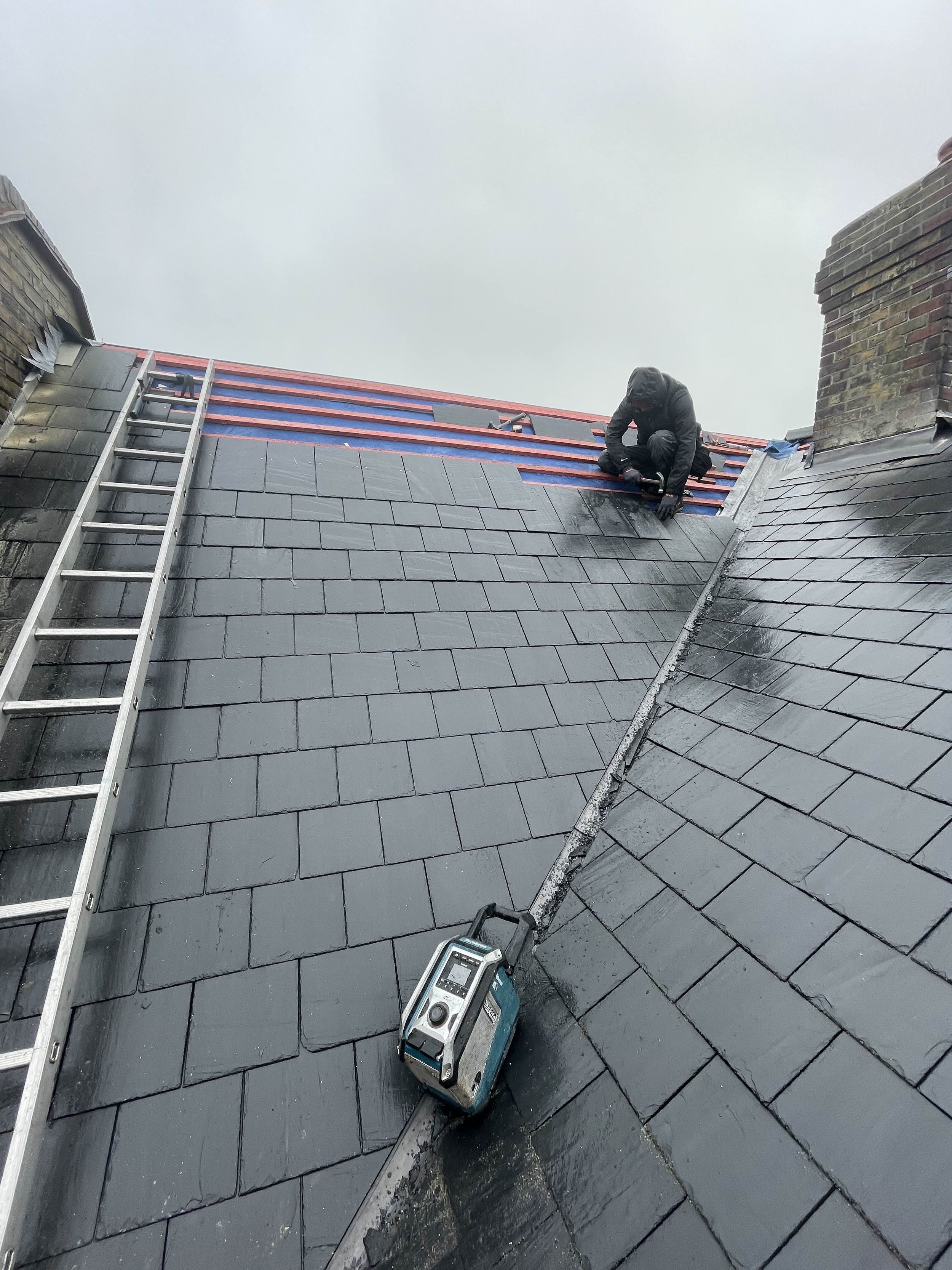 A worker in black clothing and hood working on roof shingles on a gray slate roof, with a ladder leaning against the roof and a portable work light near the edge.