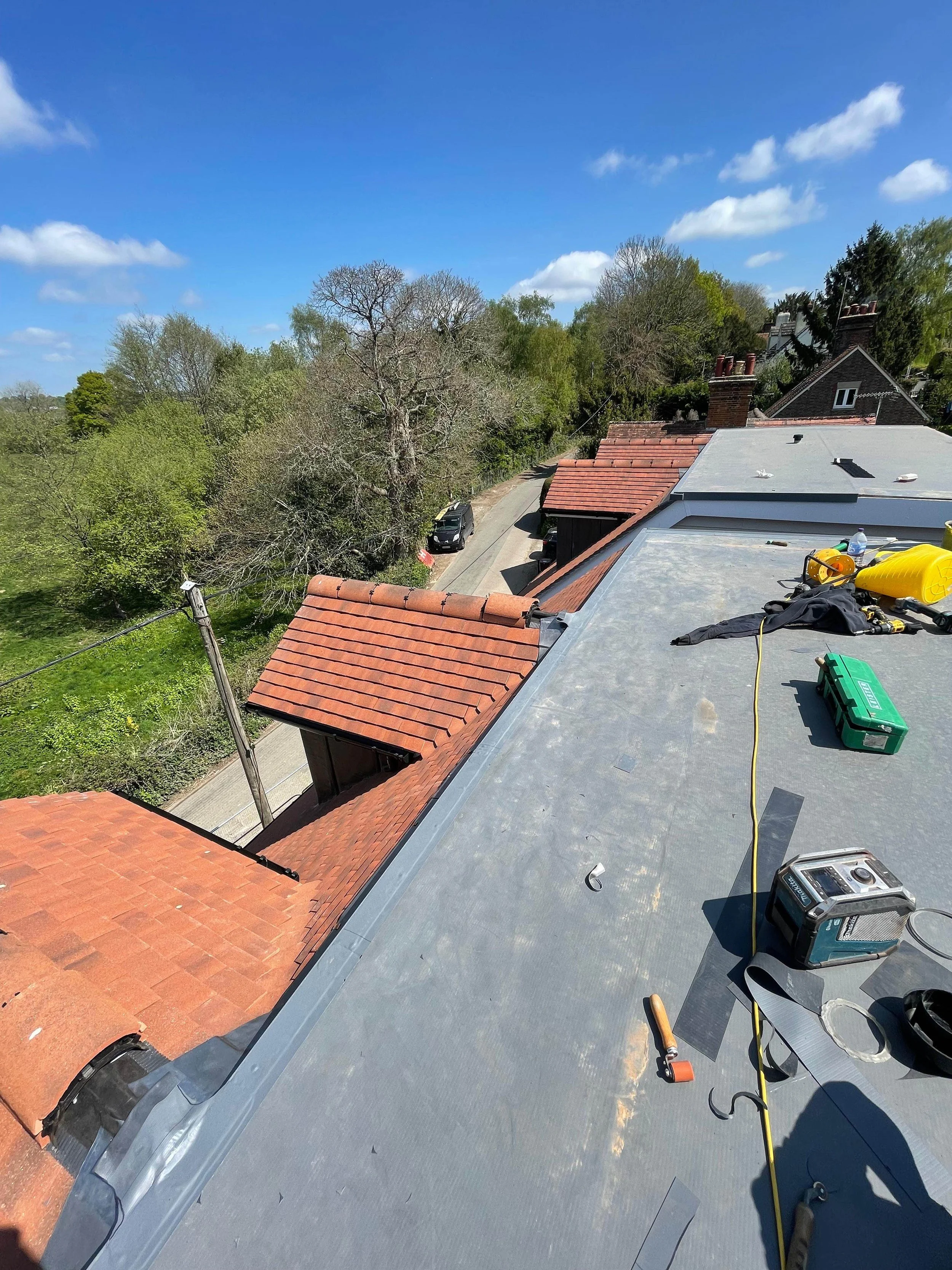 View from a rooftop showing roofing tools and equipment, neighboring rooftops with red shingles, a street, and greenery under a blue sky with clouds.