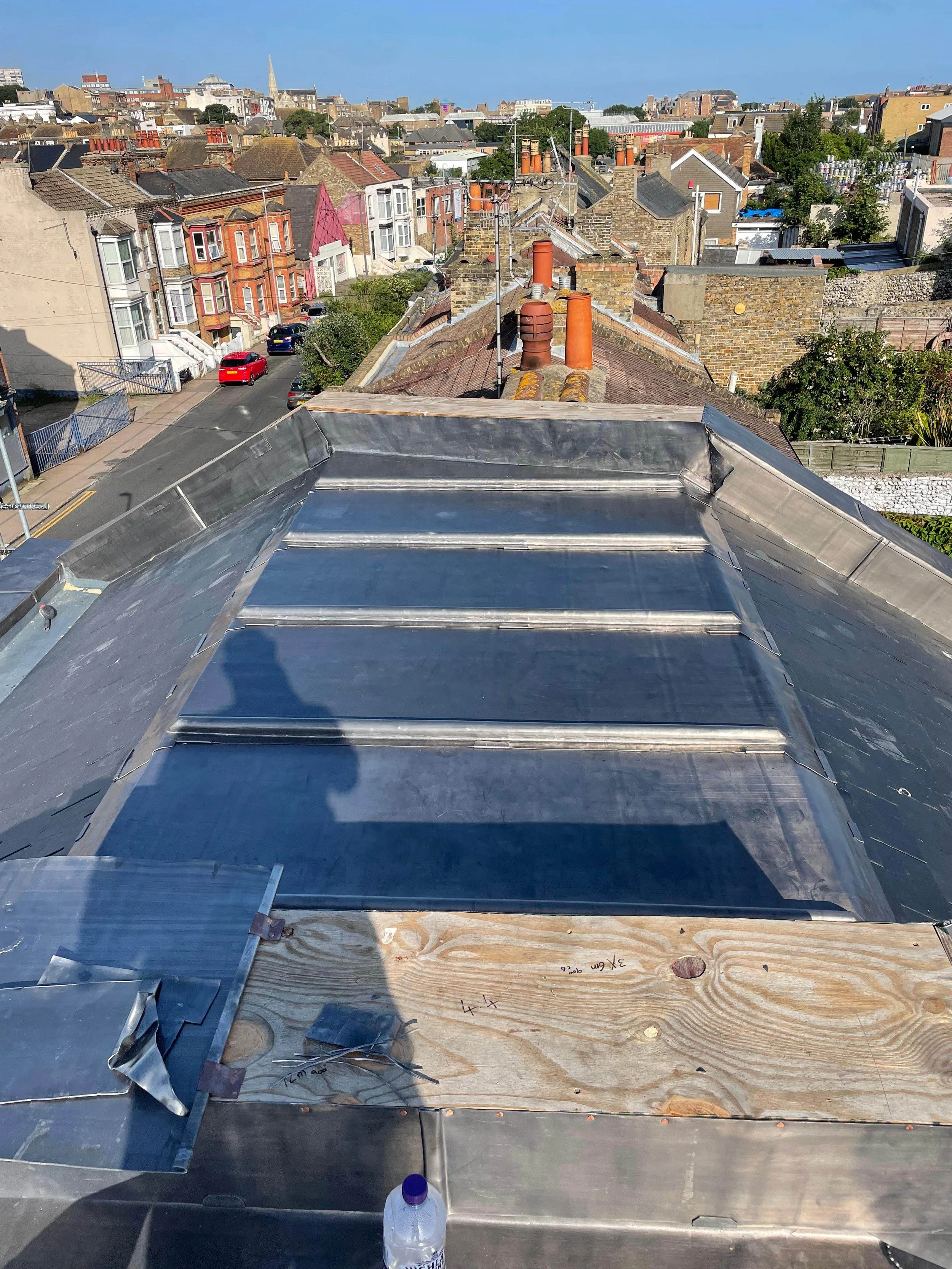 View of city rooftops with a metal roof extension in the foreground, some under construction, and a street with parked cars and colorful Victorian-style houses in the background.