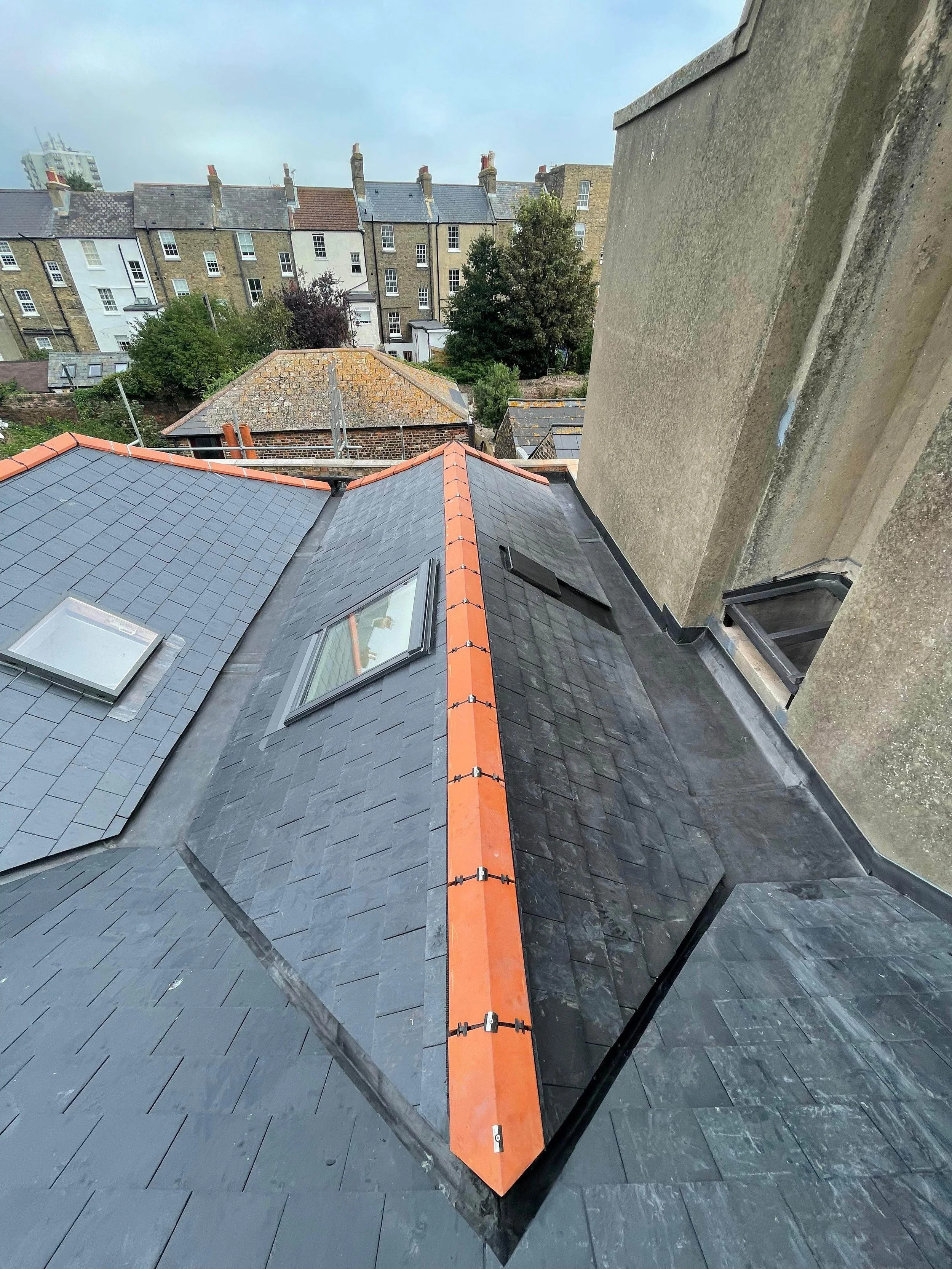 A view of a slate-tiled roof with a newly installed copper-colored ridge cap, skylights, and a chimney on the right side. In the background, there are several residential buildings, trees, and a cloudy sky.