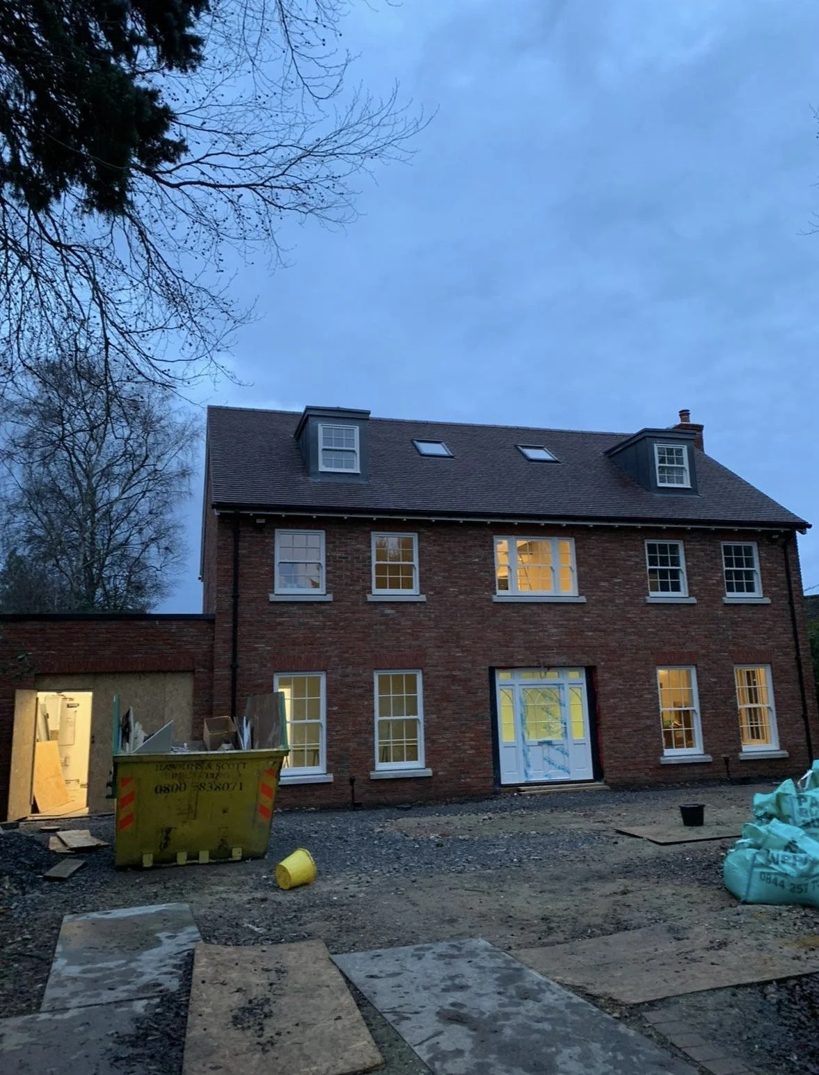 The image shows a brick house under construction during dusk, with some windows lit from inside. There is a construction site in the foreground with a yellow dumpster, construction materials, and debris scattered on the ground.
