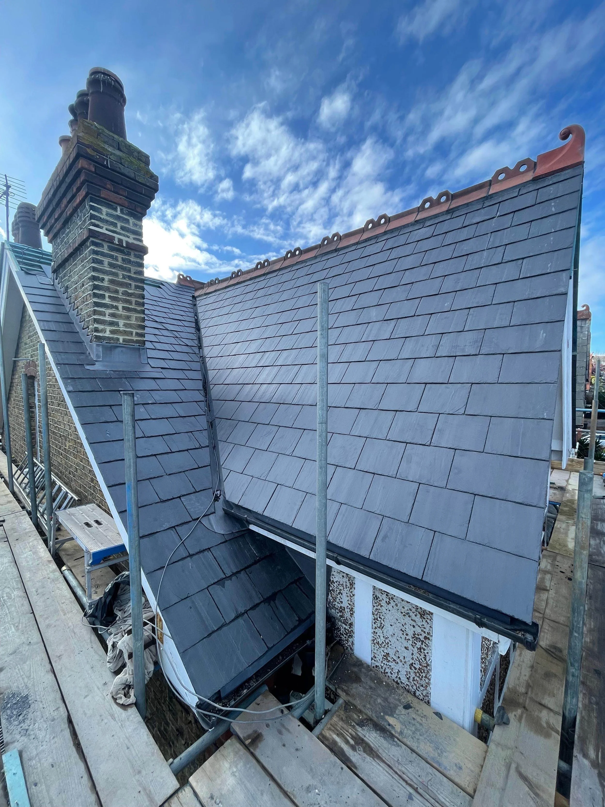 View of a house roof under construction with gray slate shingles, brick chimney with multiple stacks, scaffolding, and a cloudy sky.