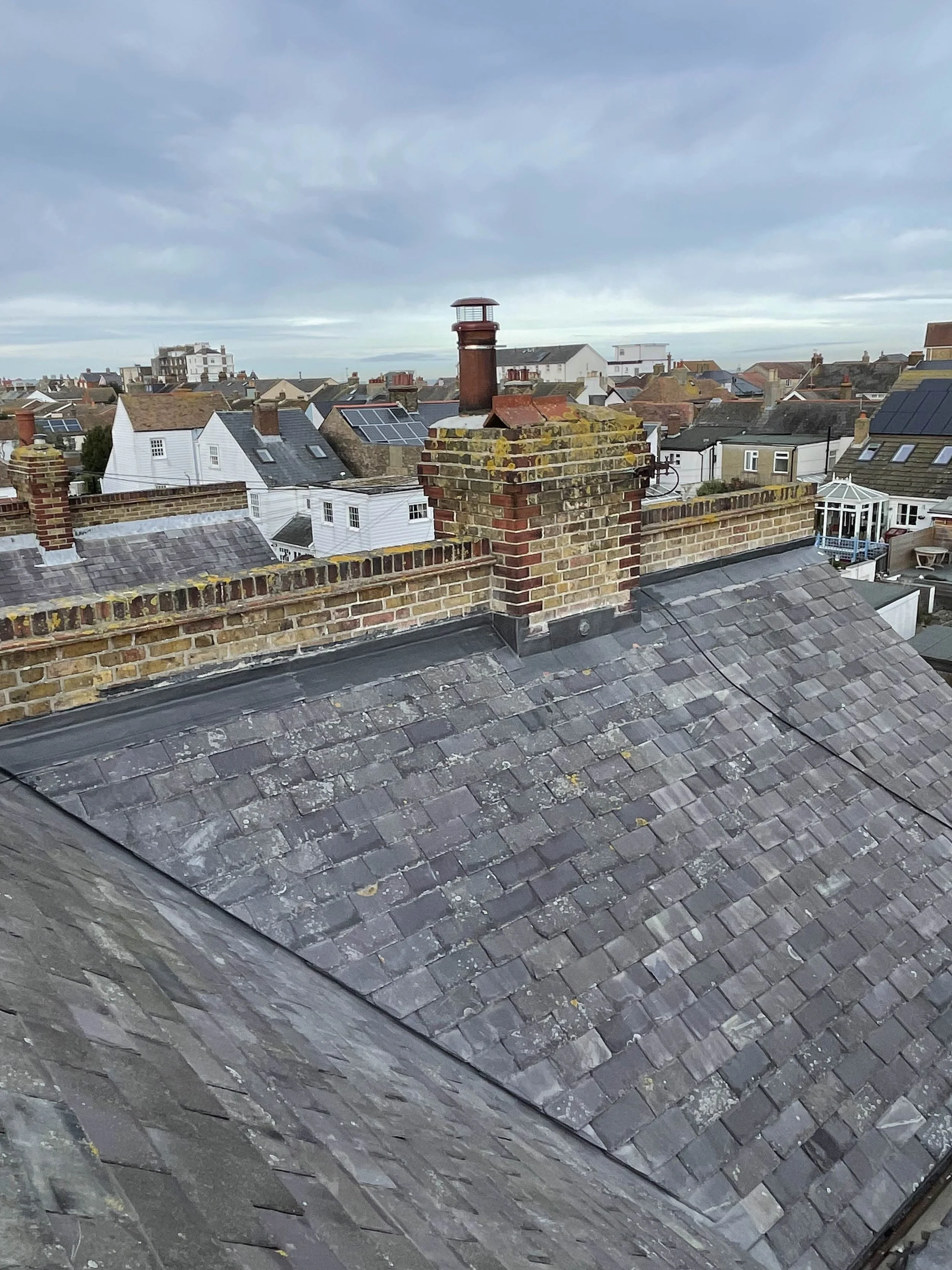 View of rooftops with a brick chimney and a small lighthouse on top of it, in a residential neighborhood under cloudy sky.