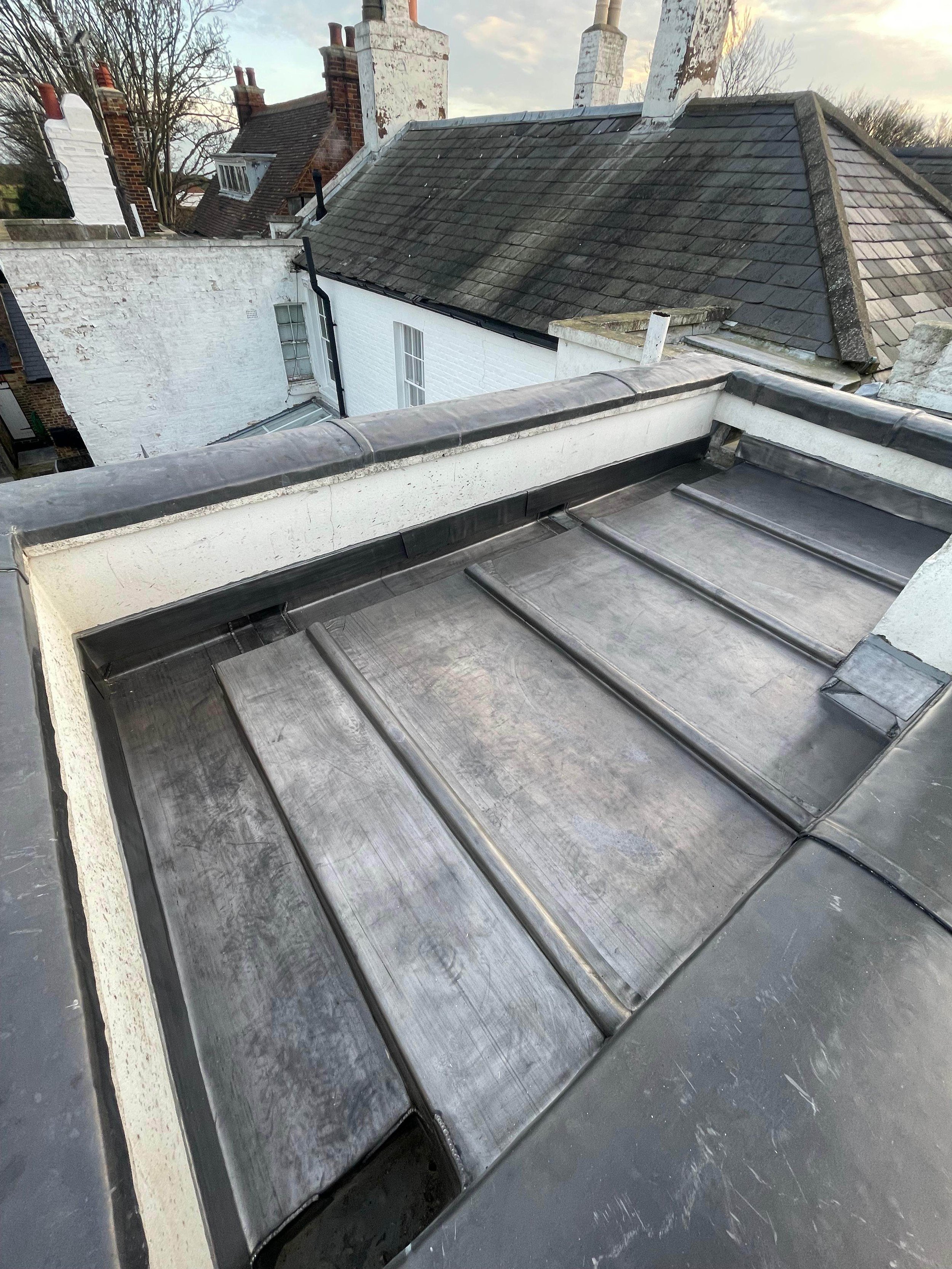 View of a metal roof on a building with neighboring rooftops and chimneys, some with brick and others with white painted surfaces, under a cloudy sky.