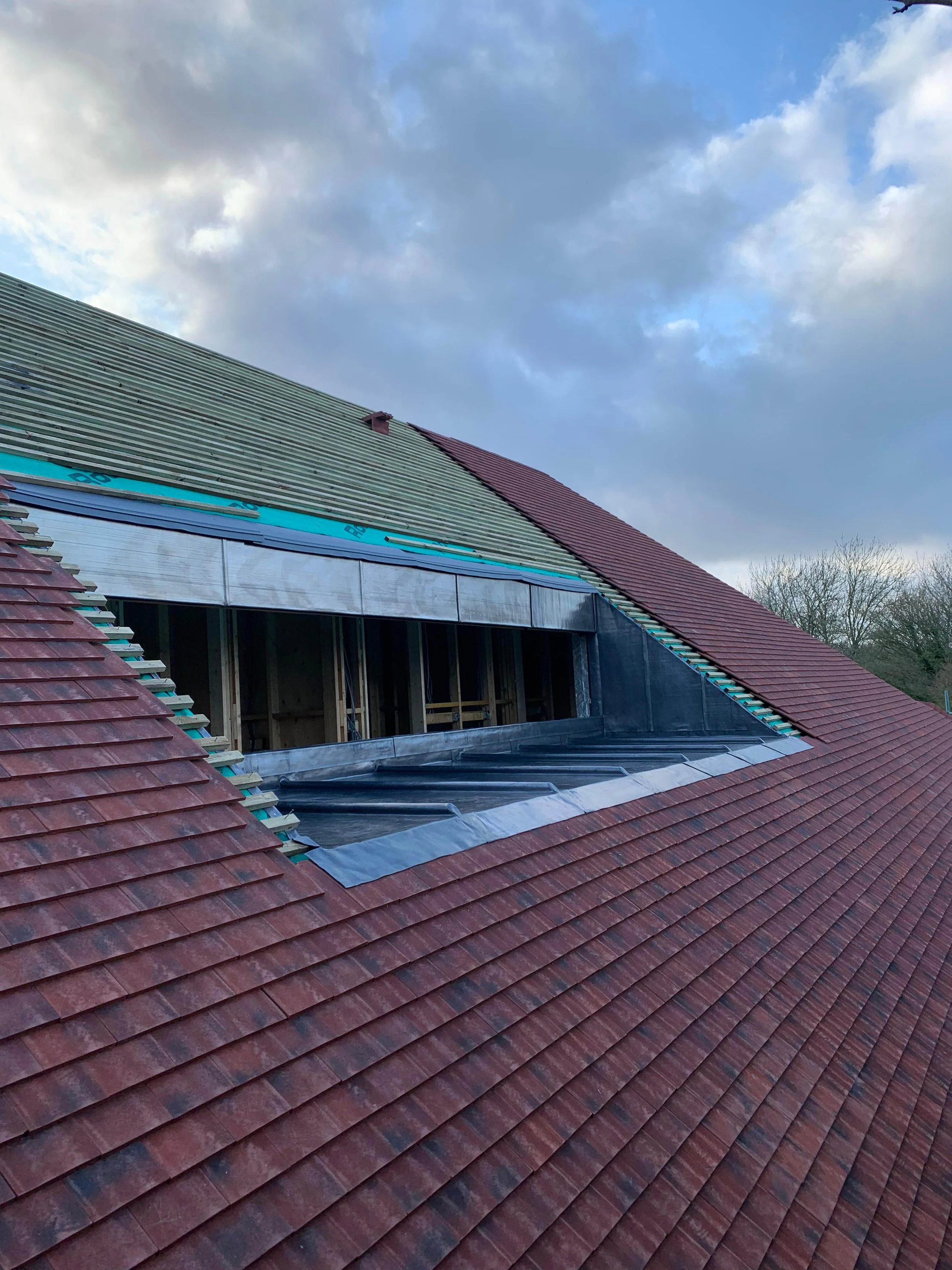 Under construction roof with partially installed roofing materials, including red roof tiles and a skylight opening, on a building under cloudy sky.