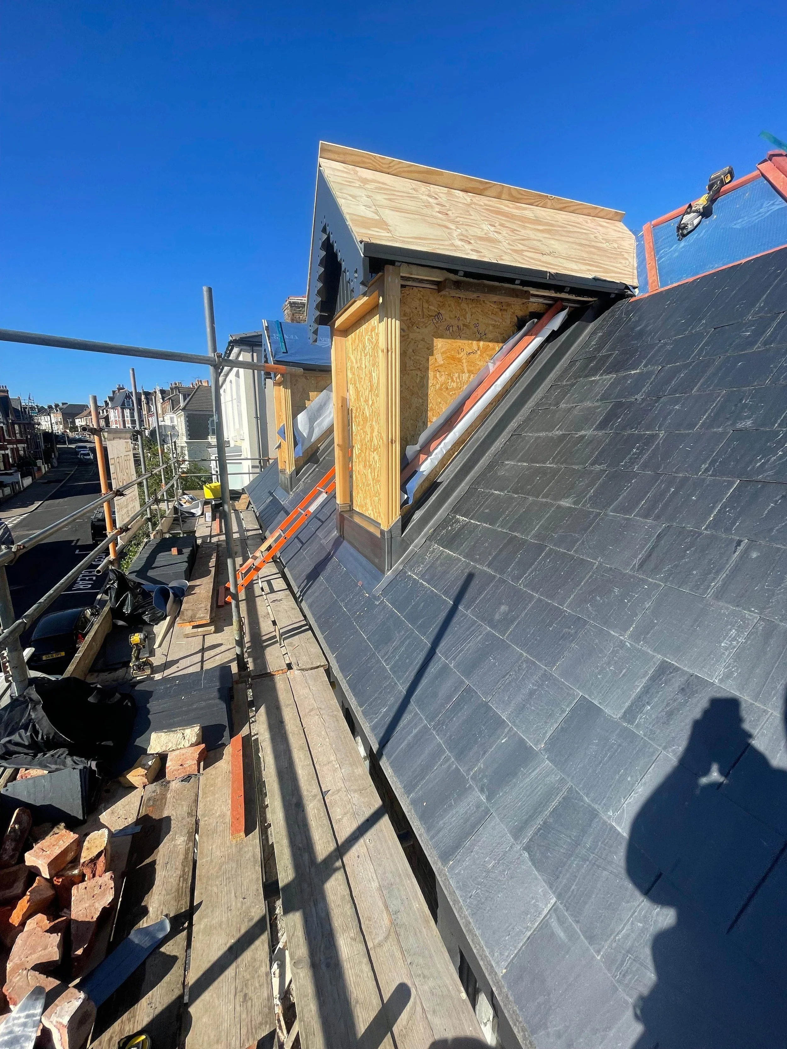 Construction site on a roof with scaffolding, showing a new dormer window being built with wood framing and black roofing tiles under clear blue sky.