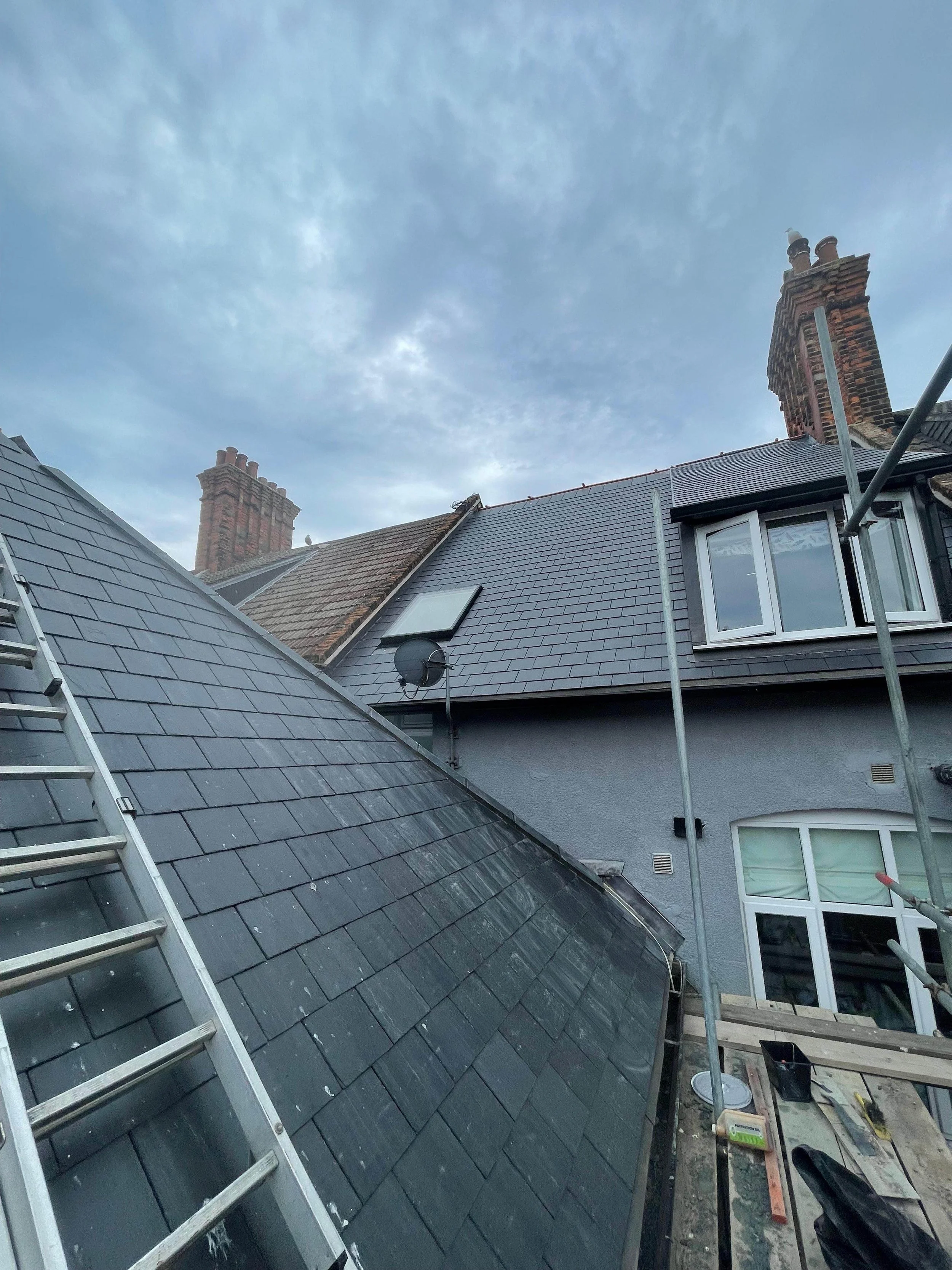 View of residential rooftops with gray tiles, chimney stacks, a satellite dish, work scaffolding, and an aluminum ladder against the roof on a cloudy day.