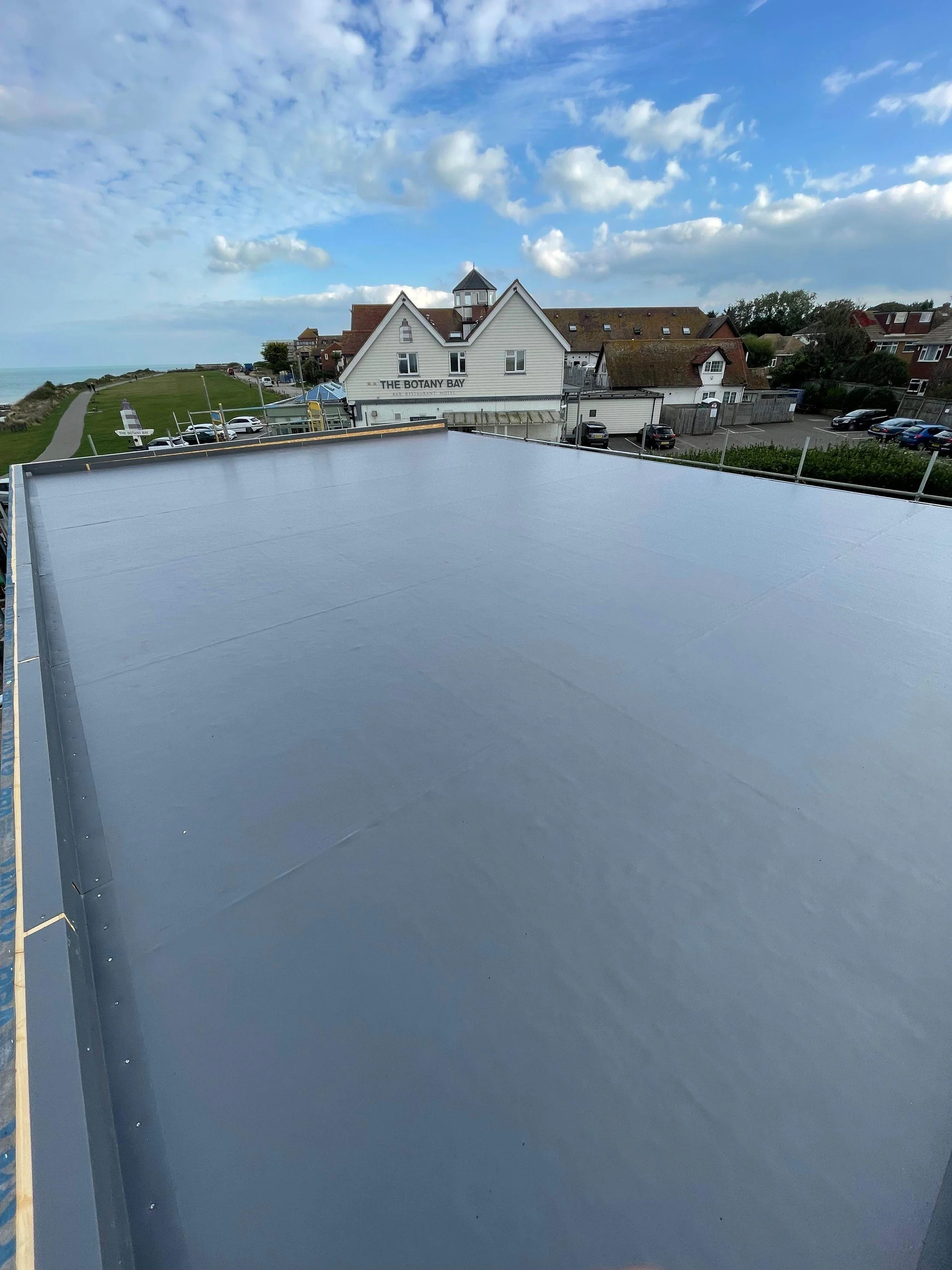 View from a rooftop with a grey surface, looking toward a coastal town with houses and a café named 'The Botany Bay' under a partly cloudy sky.