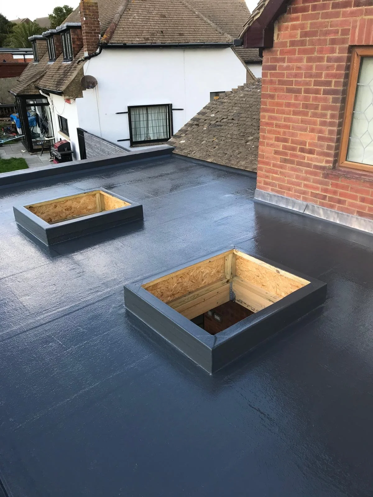 A flat roof with two square skylights, one of which is open, showing wood framing inside; the roof is coated with black waterproof membrane.