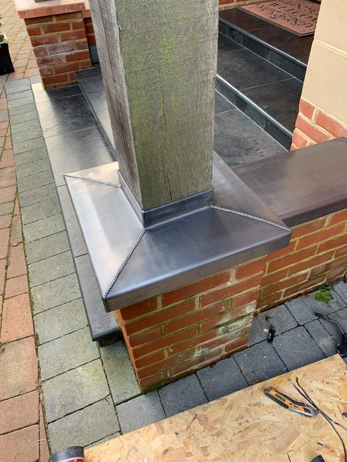 A close-up of a brick pillar with a vertical wooden post attached to a metal base, part of an outdoor structure under construction. There are tools and construction materials around.