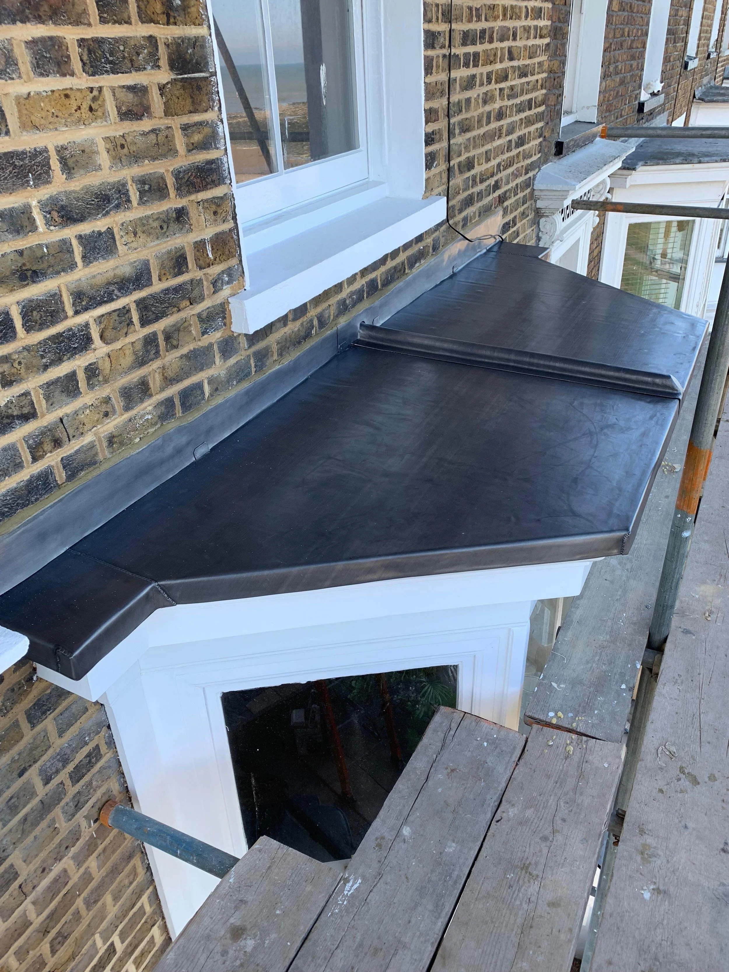Close-up of a building's exterior, showing a brick wall, a window frame, and a black protective cover on a small roof extension, with scaffolding nearby.