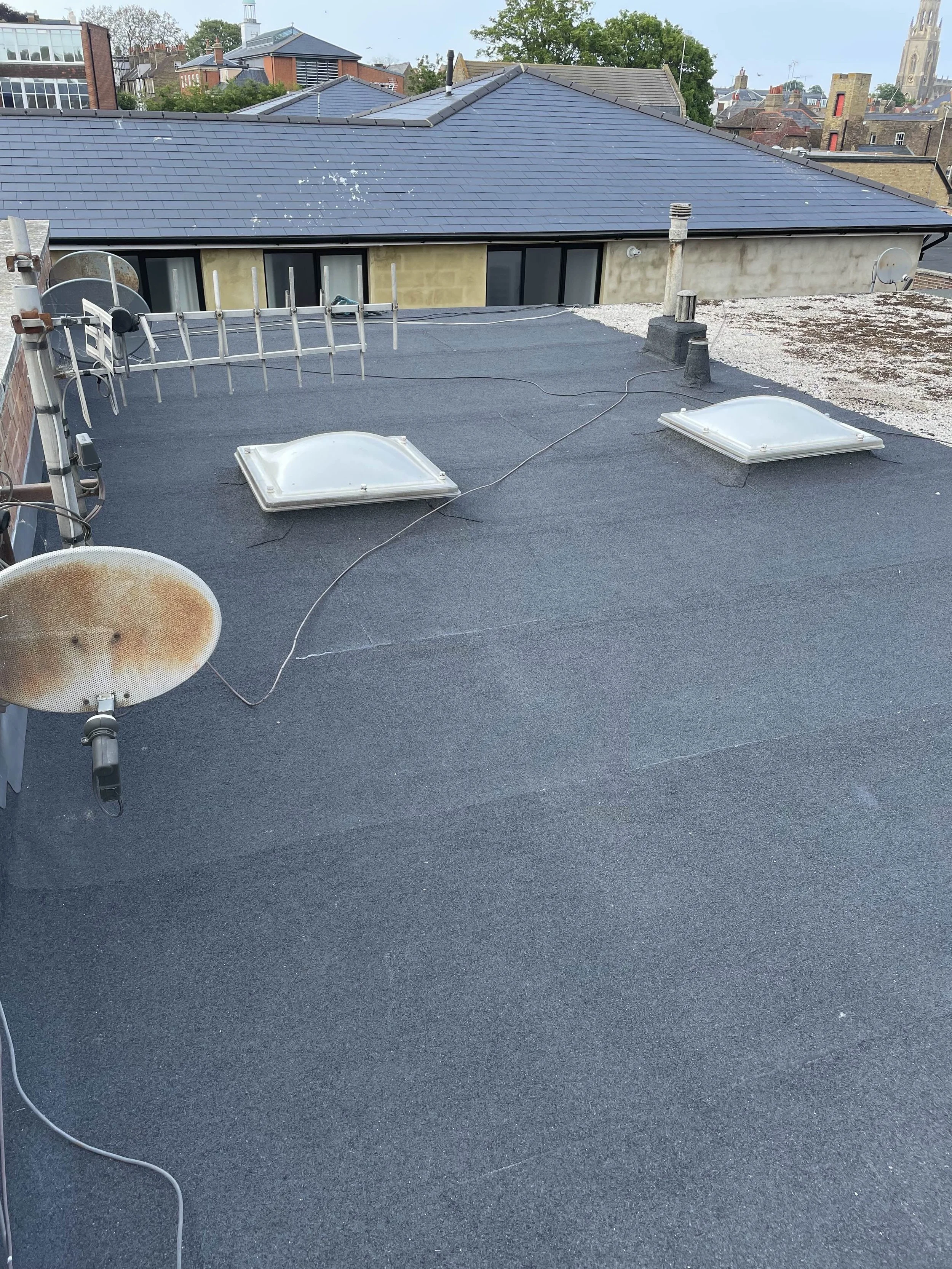 Rooftop with black weatherproof surface, two skylights, aerial antenna, and various satellite dishes, with neighboring buildings and a church in the background.