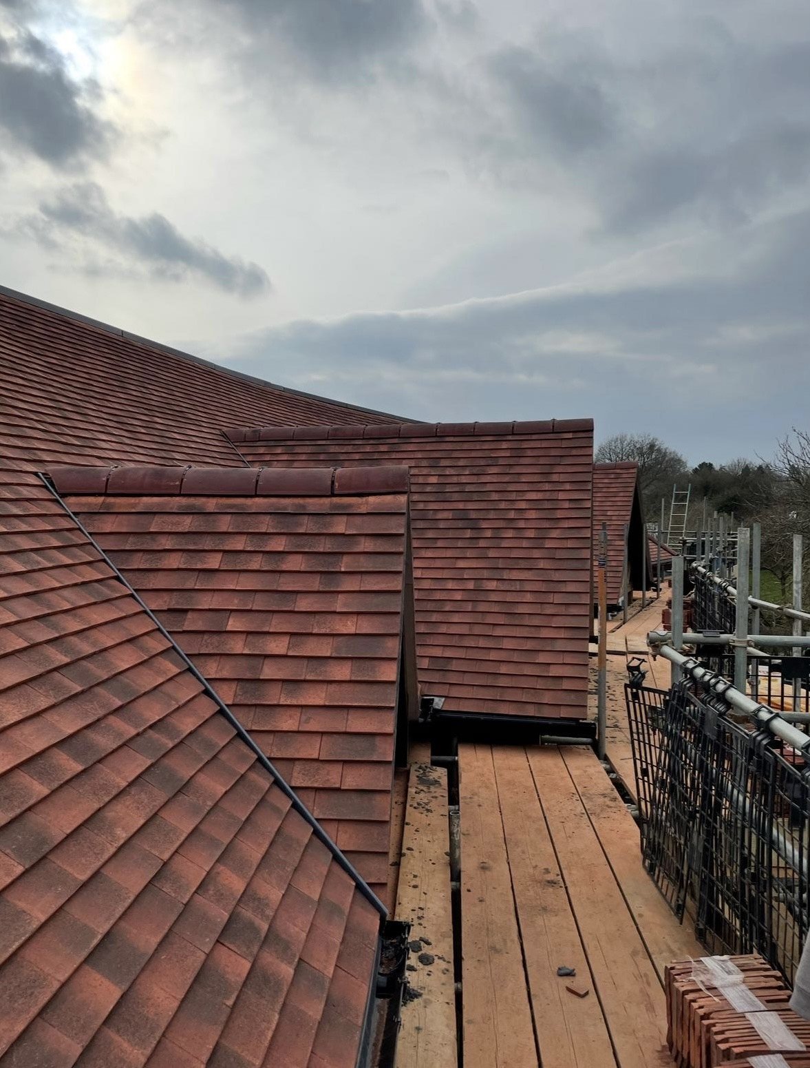 Red tiled roof sections under a cloudy sky, with scaffolding and building materials on a wooden walkway, suggesting construction or maintenance work.