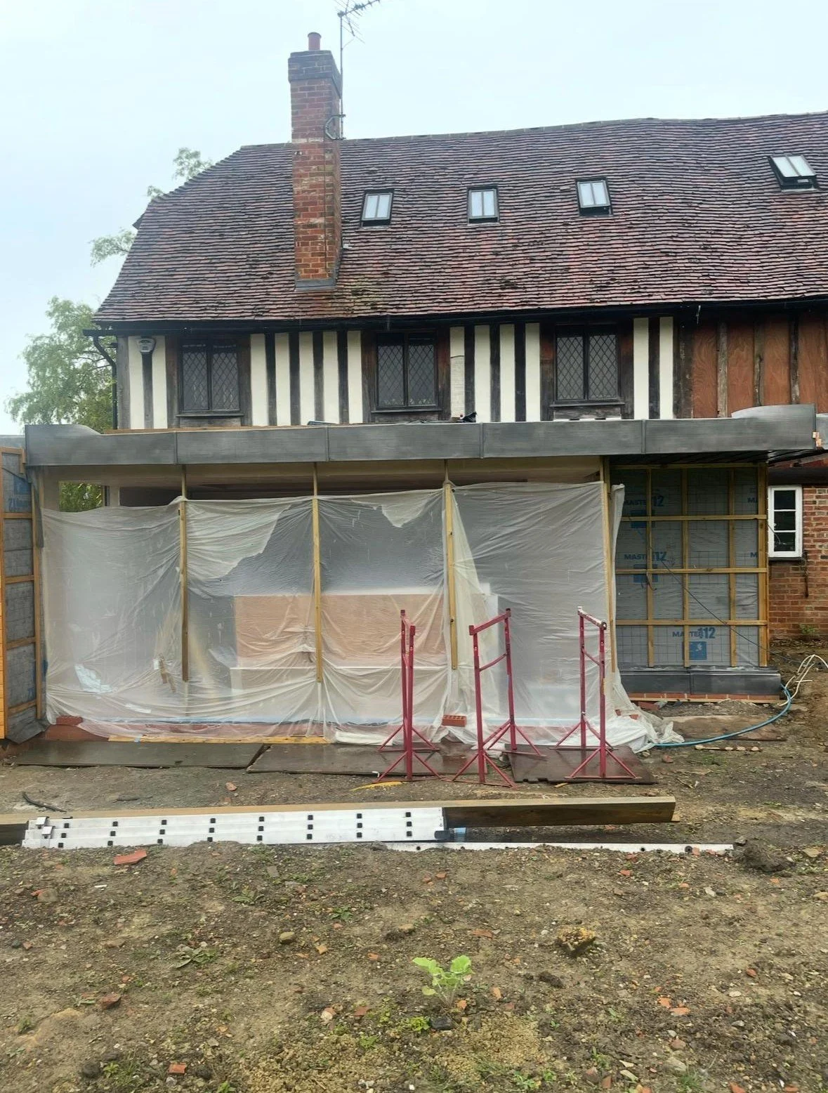 Under construction extension of a brick house with a wooden-frame extension, covered in plastic sheeting, in a garden area with construction materials and barriers.