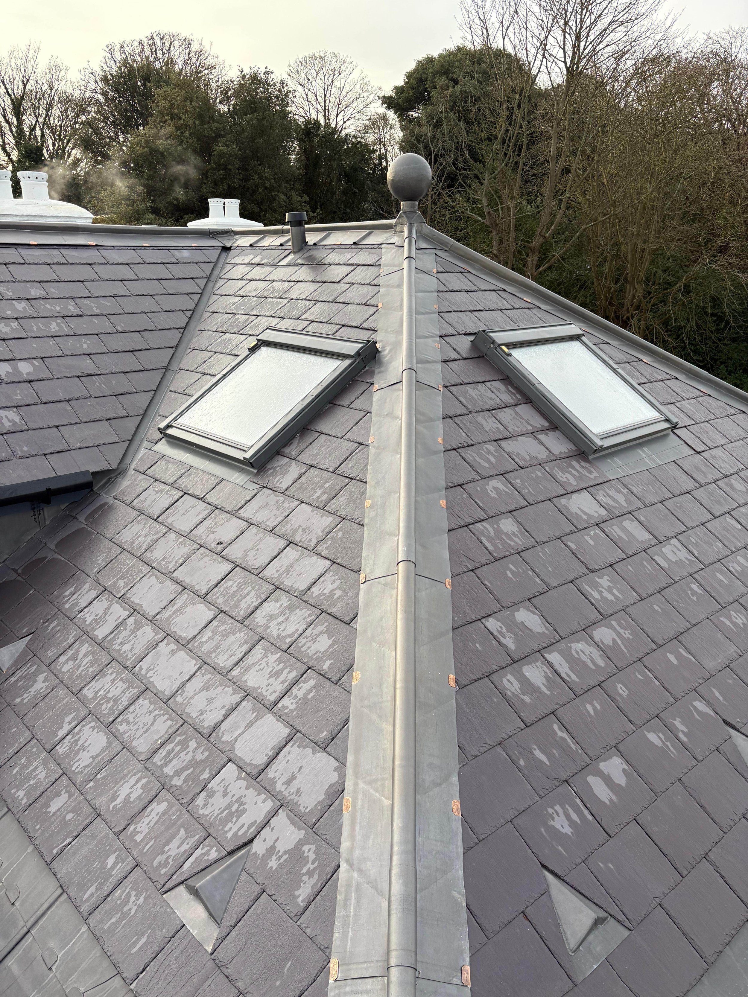 Roof with shingles, two skylights, a metal ridge cap, roof vents, and a ball-shaped finial, surrounded by trees.