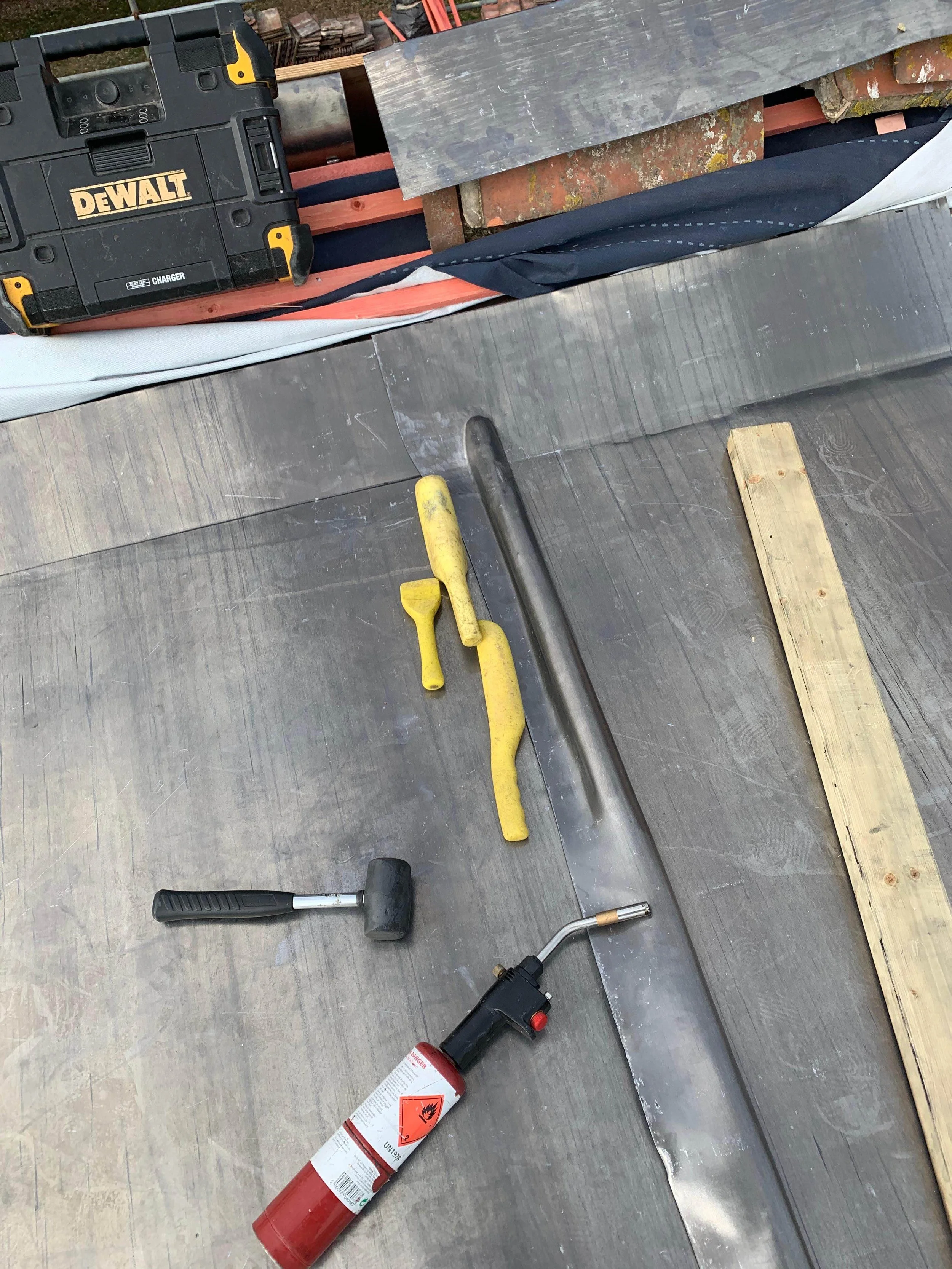Tools and materials for construction work, including a yellow hammer, a metal pipe, a propane torch, a wooden plank, and a DeWalt cordless drill, are arranged on a wooden floor near a construction site with brick and wood materials.