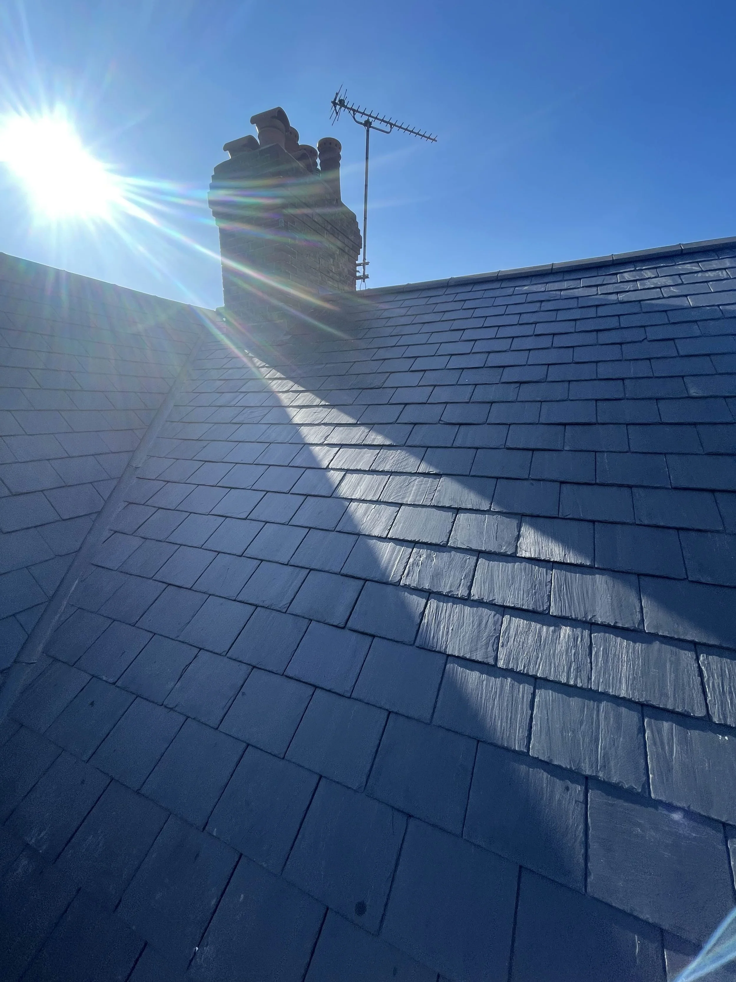 Sun shining brightly over a rooftop with gray slate shingles and a chimney with terracotta chimney pots.