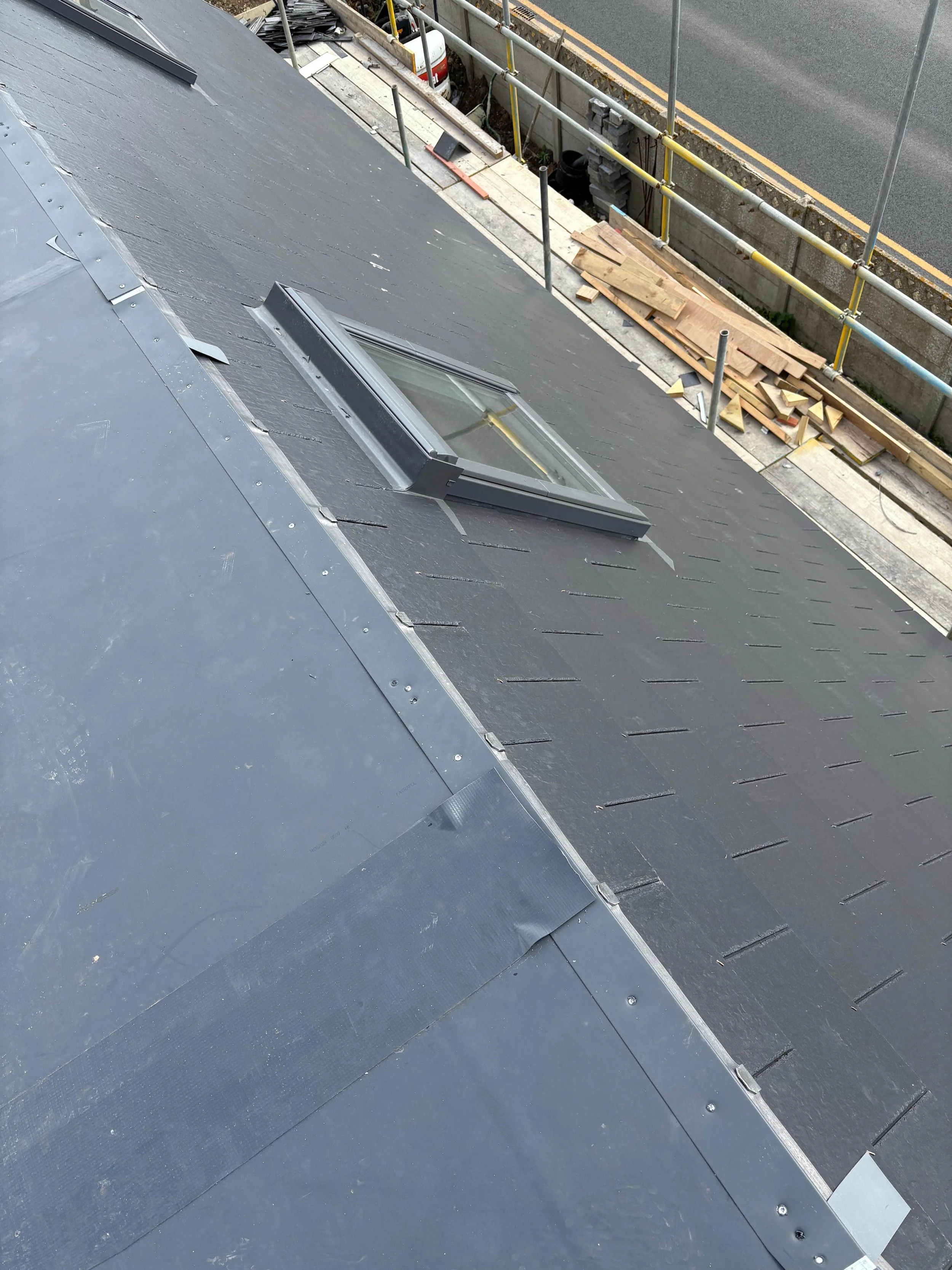 A rooftop with a skylight window under construction, showing roofing materials and scaffolding along the edge of the roof.