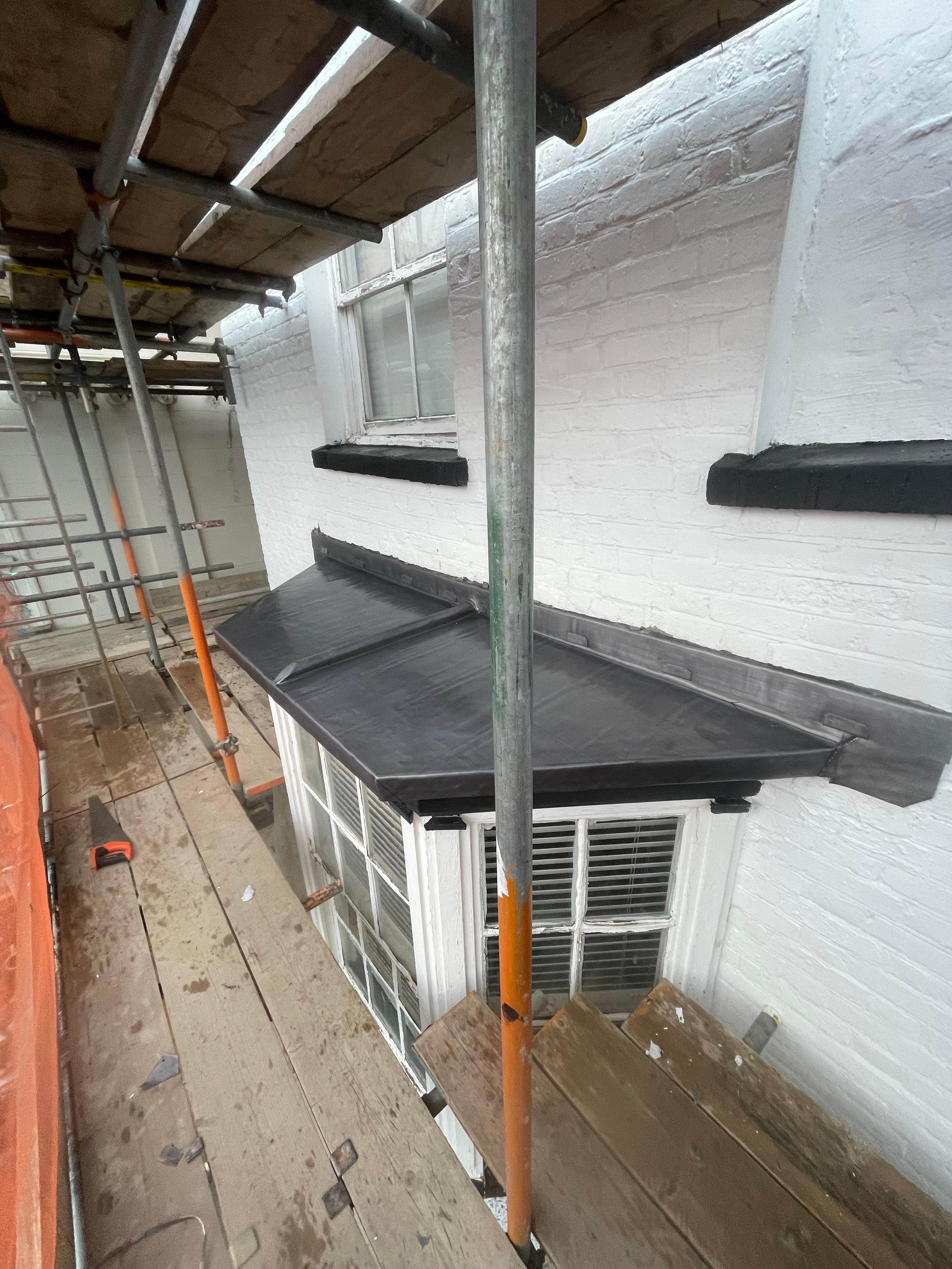 Construction scaffolding around a house with a window, showing a new black-roofed awning above the window and some wet wooden planks on the scaffold platform.