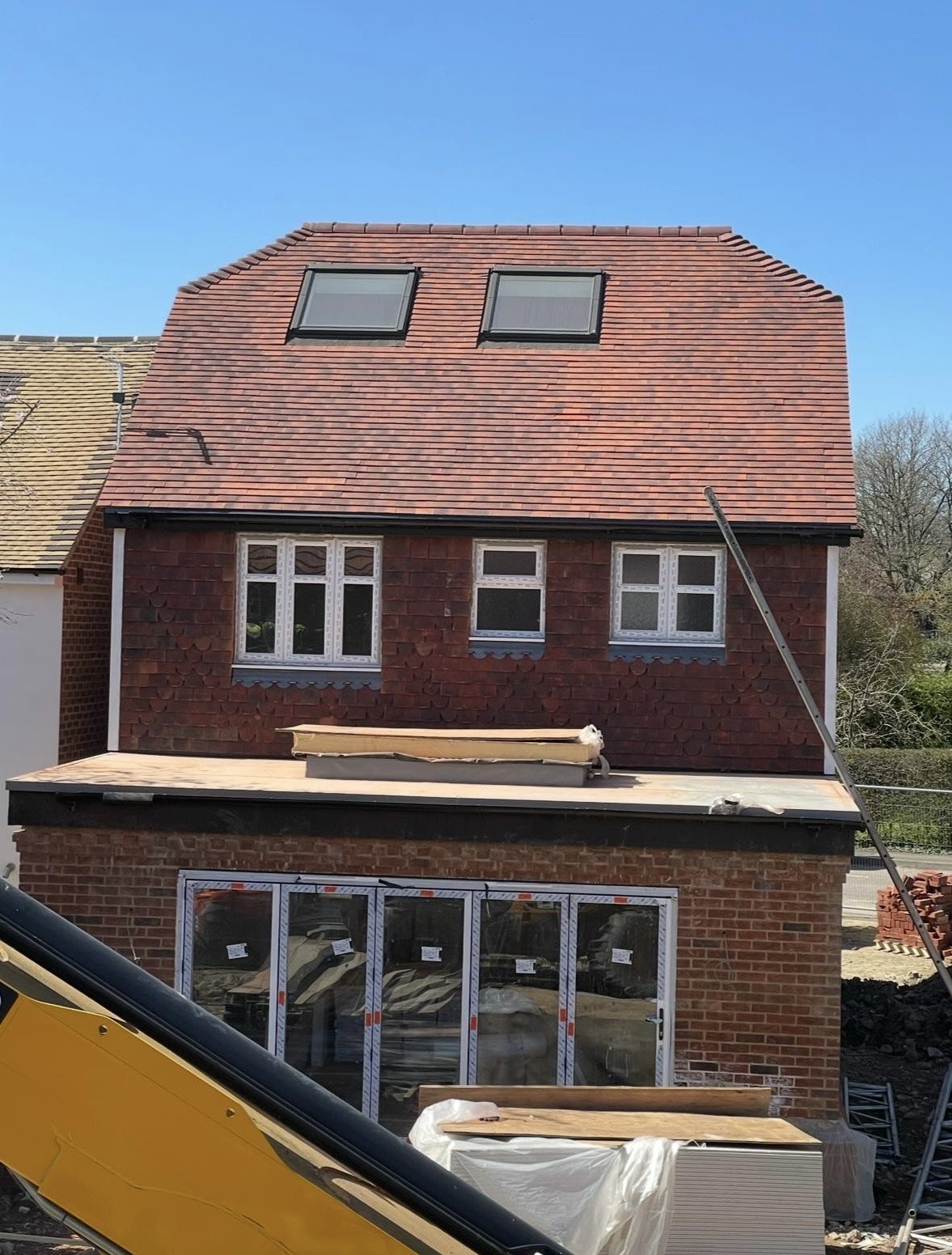 Front view of a house under construction with a red brick exterior and multiple windows, including two large skylights on the roof, and construction materials and equipment visible in the foreground.
