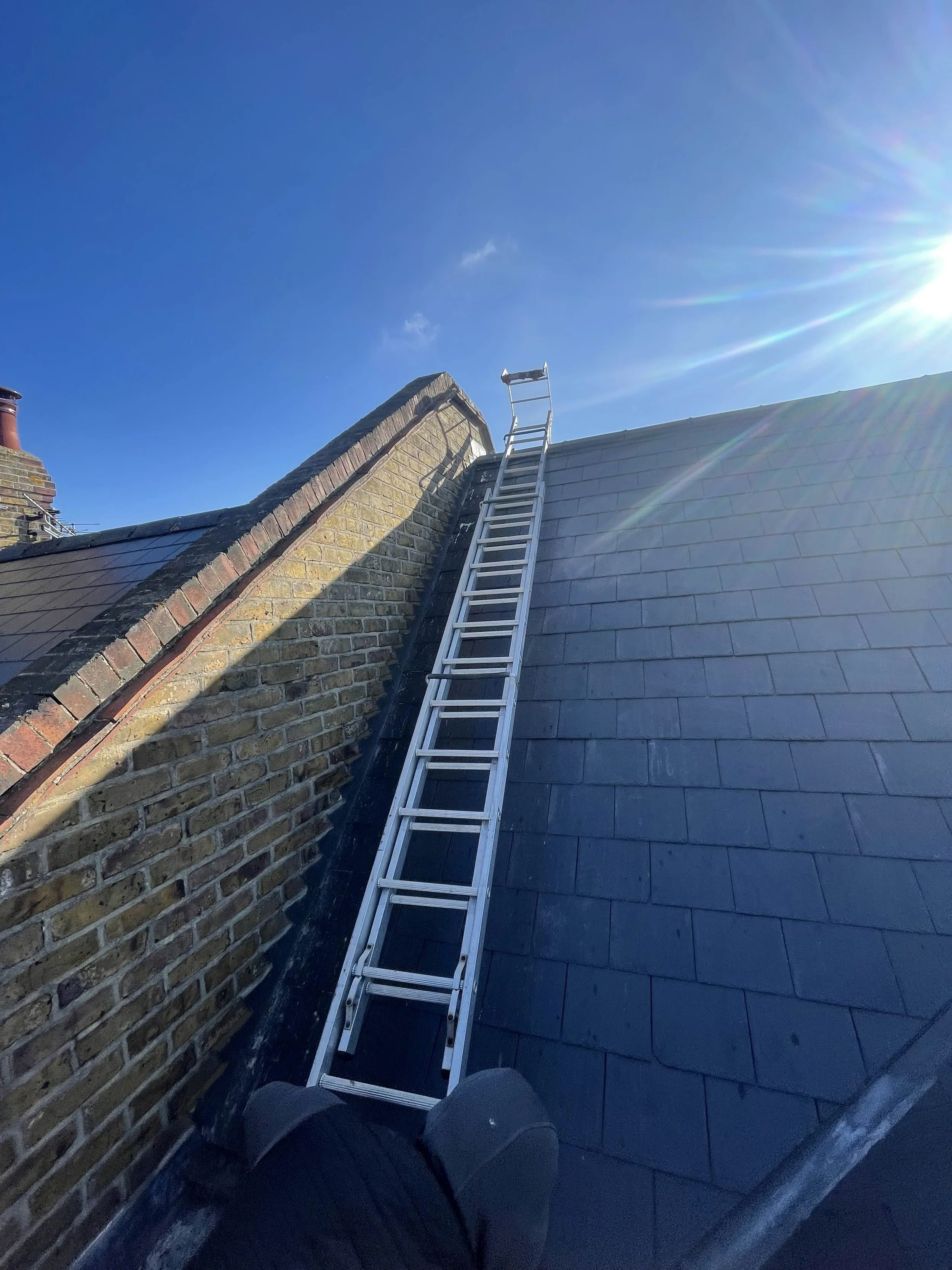 Looking up a roof with a ladder leaning against it and a person's foot visible in the foreground, under a clear blue sky with the sun shining.
