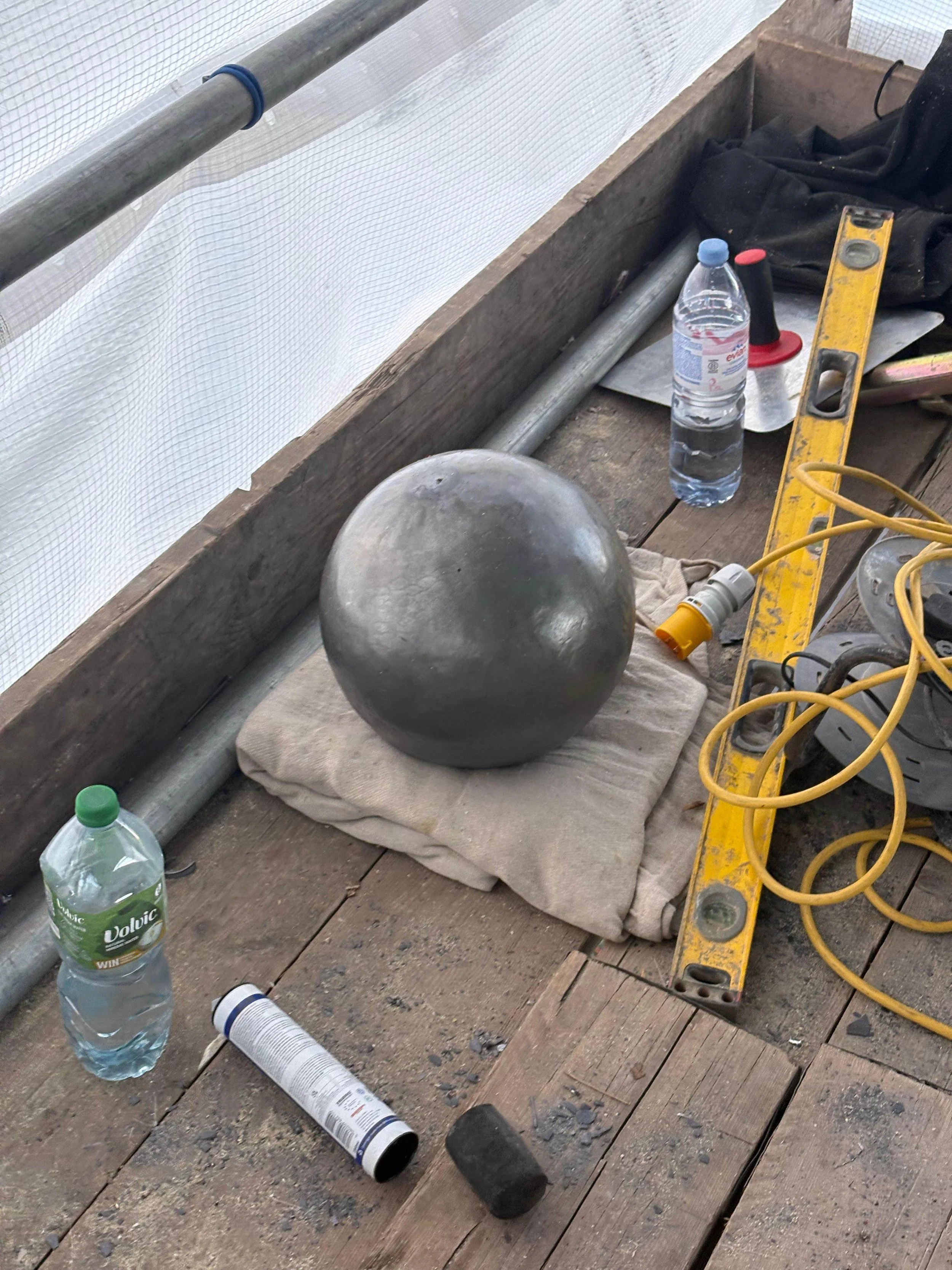 A construction site with a metallic sphere on a cloth, water bottles, a level, various tools, an extension cord, and construction debris on a wooden floor.