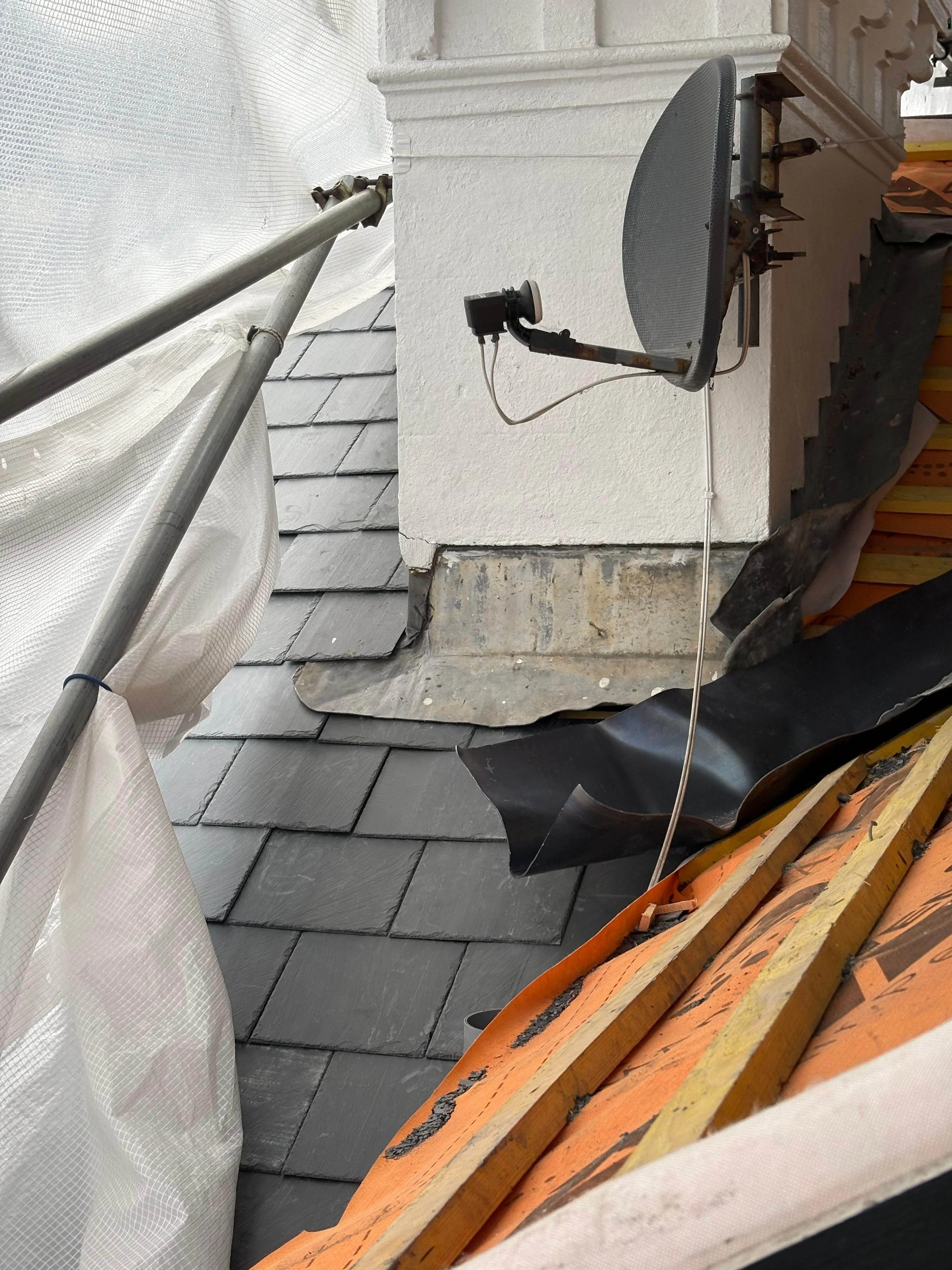 Roof construction scene with slate tiles, scaffolding, and a satellite dish attached to a white wall.