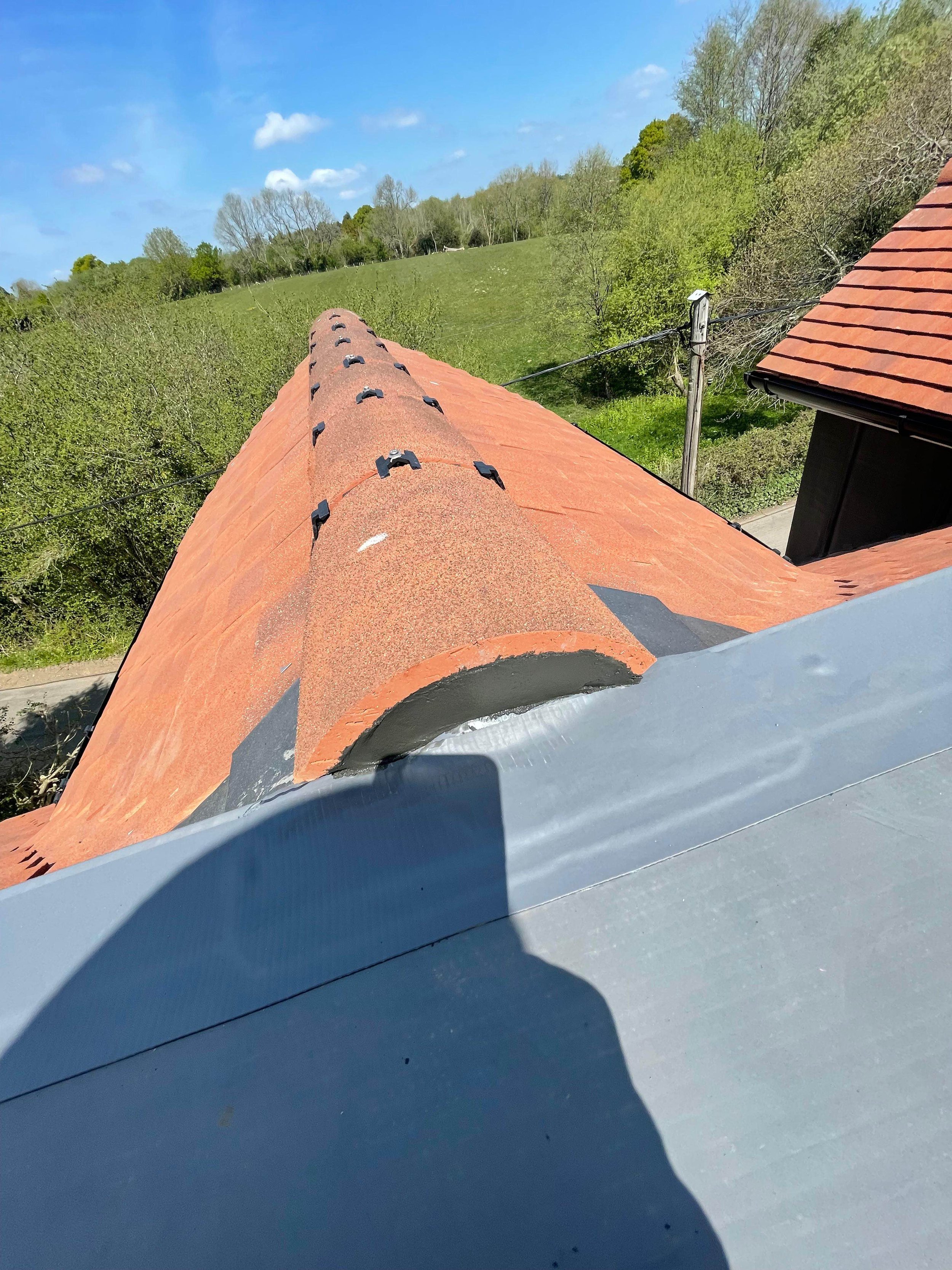 View of a rooftop with orange shingles extending into a green landscape with trees and blue sky.