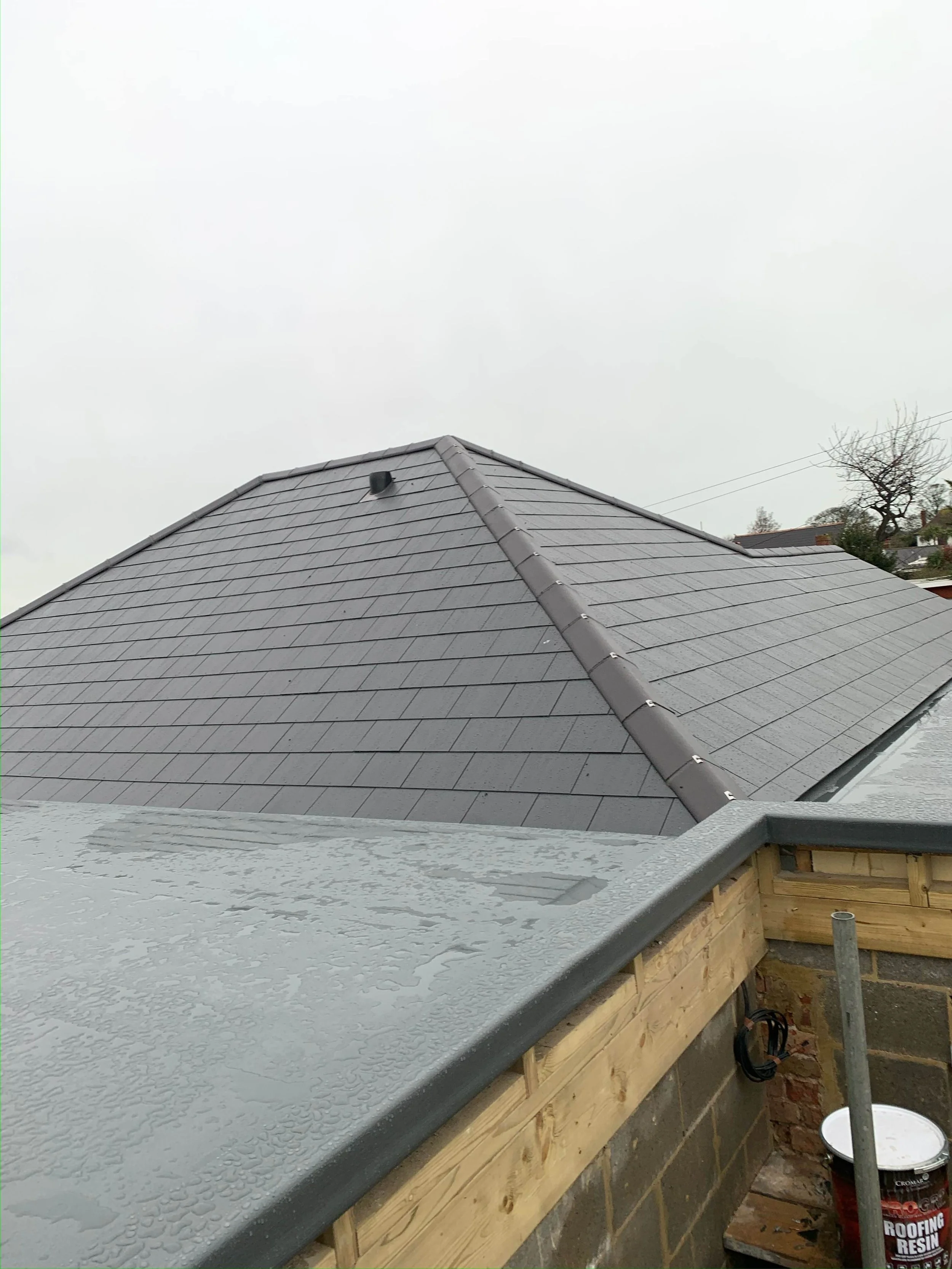 View of a house roof with newly installed gray shingles and a rain-soaked ledge in the foreground, part of the roof under construction with visible brick and wood framing.