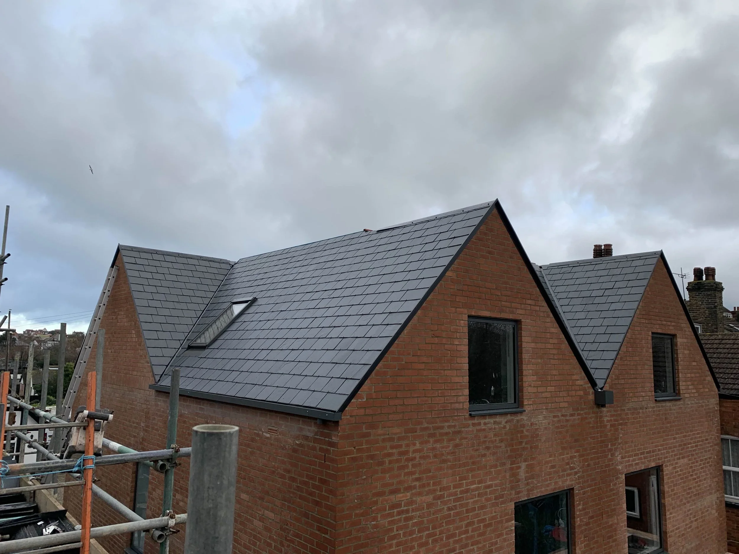 A brick house with newly installed dark grey slate roof, skylight window, and surrounding cloudy sky.