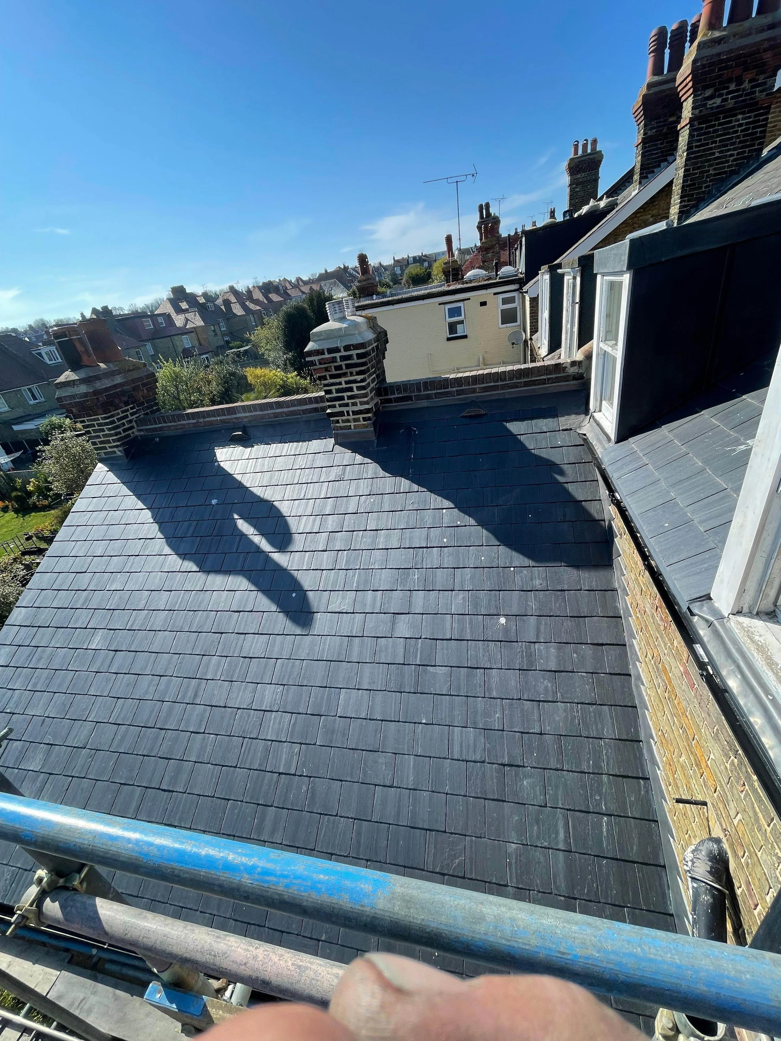 View of a rooftop with black slate tiles, chimney stacks, and surrounding residential buildings under a blue sky.