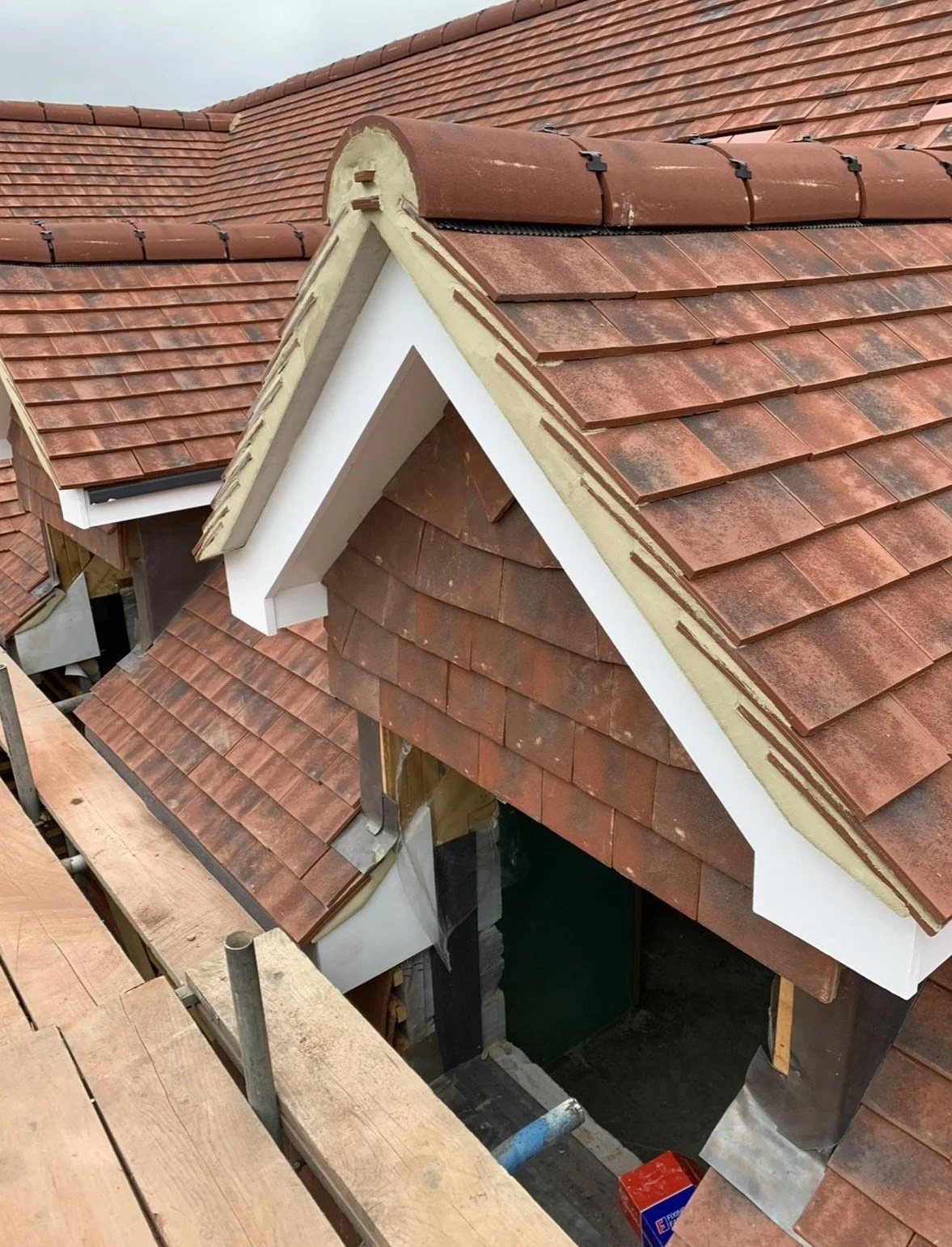 Construction site showing a building with a complex roof structure, red clay tiles, and white trim, with scaffolding nearby.