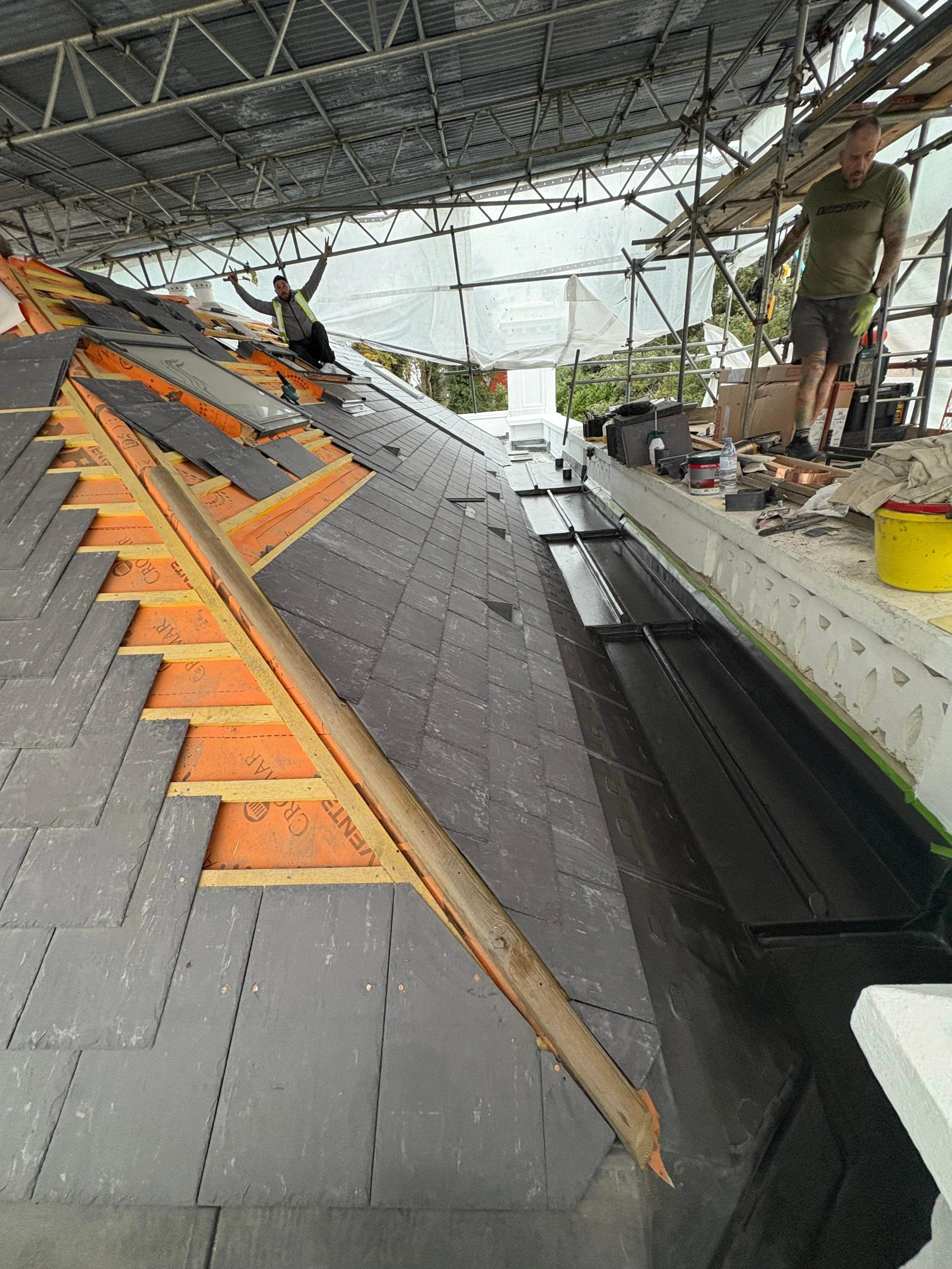 Roof construction with shingles being installed; two workers are on the roof with tools and materials, working under a temporary covering.