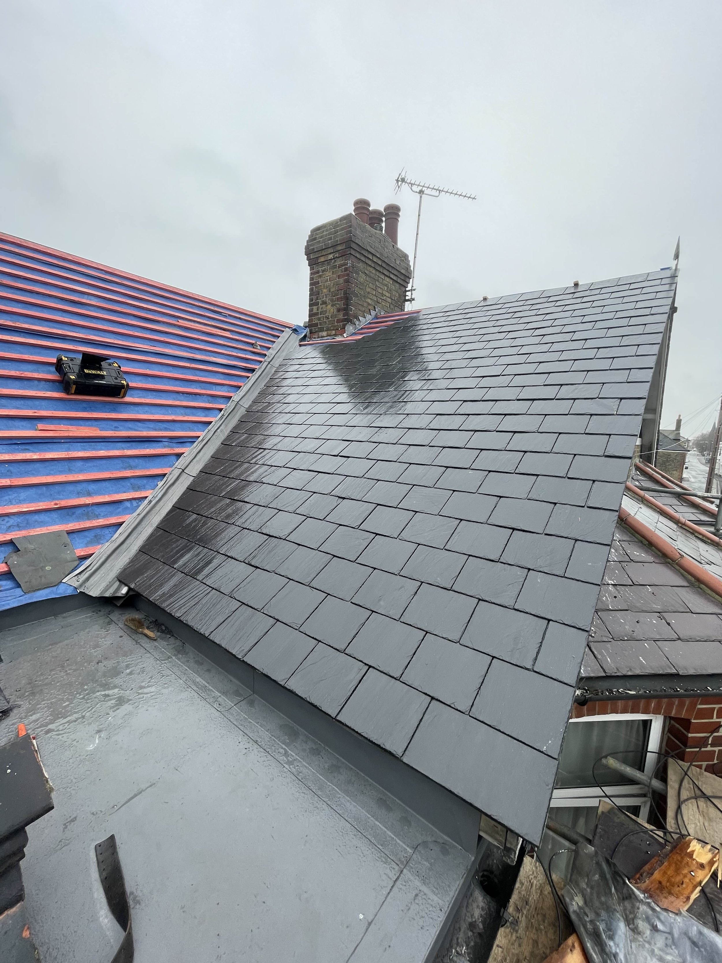 Close-up view of a house roof with new dark grey slate tiles, adjacent to an older roof with blue and red tiles.