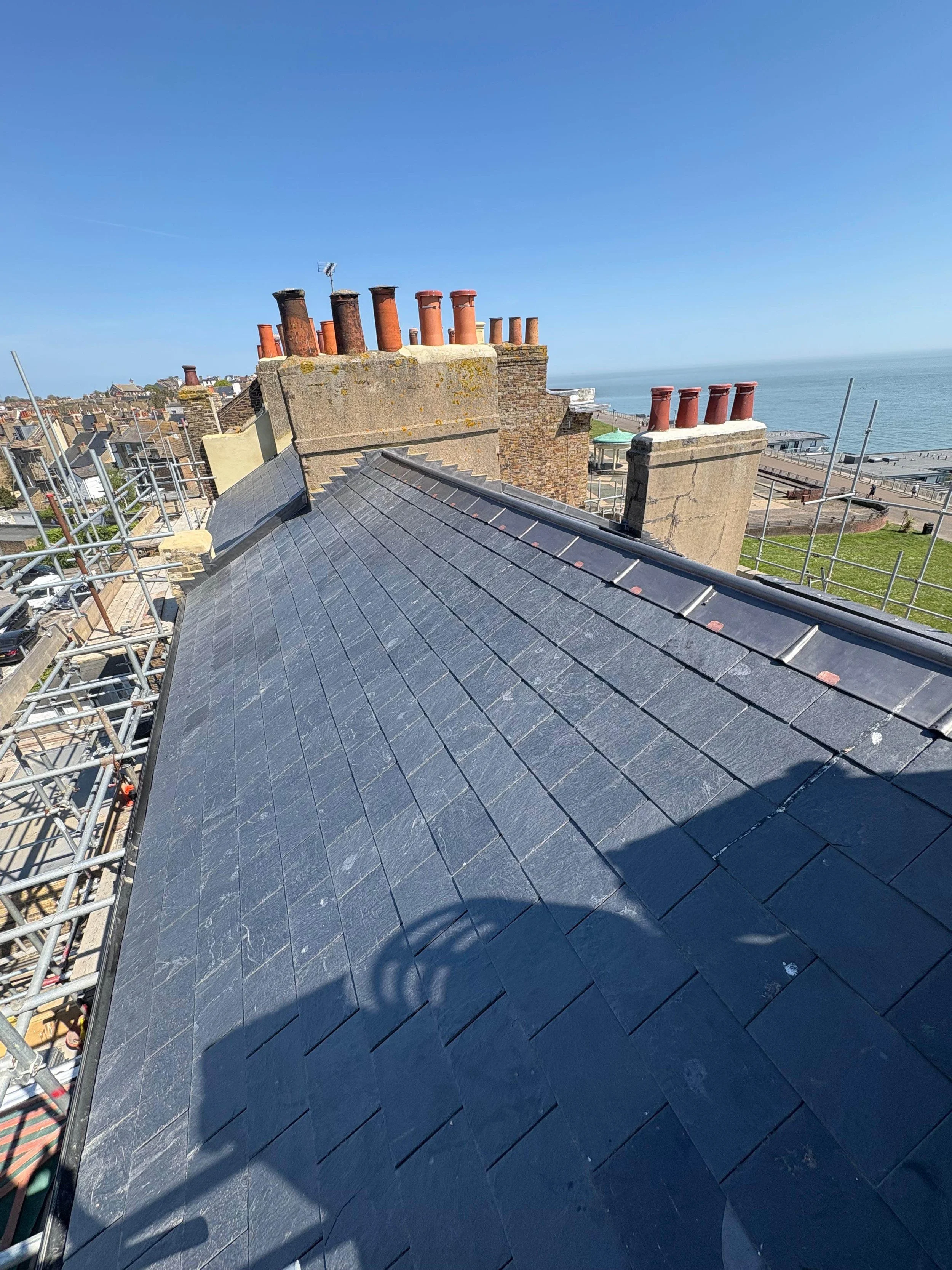 View of a dark gray slate roof with multiple brick chimneys on a building, overlooking scaffolding, neighboring buildings, the ocean, and clear blue sky.