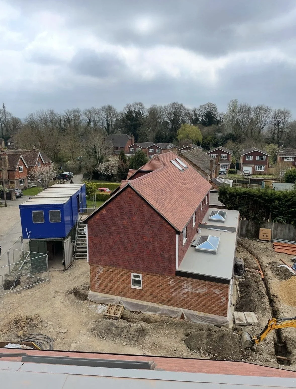 Construction site view of a brick building with a red tiled roof, two skylights, and a blue temporary office container, with construction equipment and dirt surrounding it.