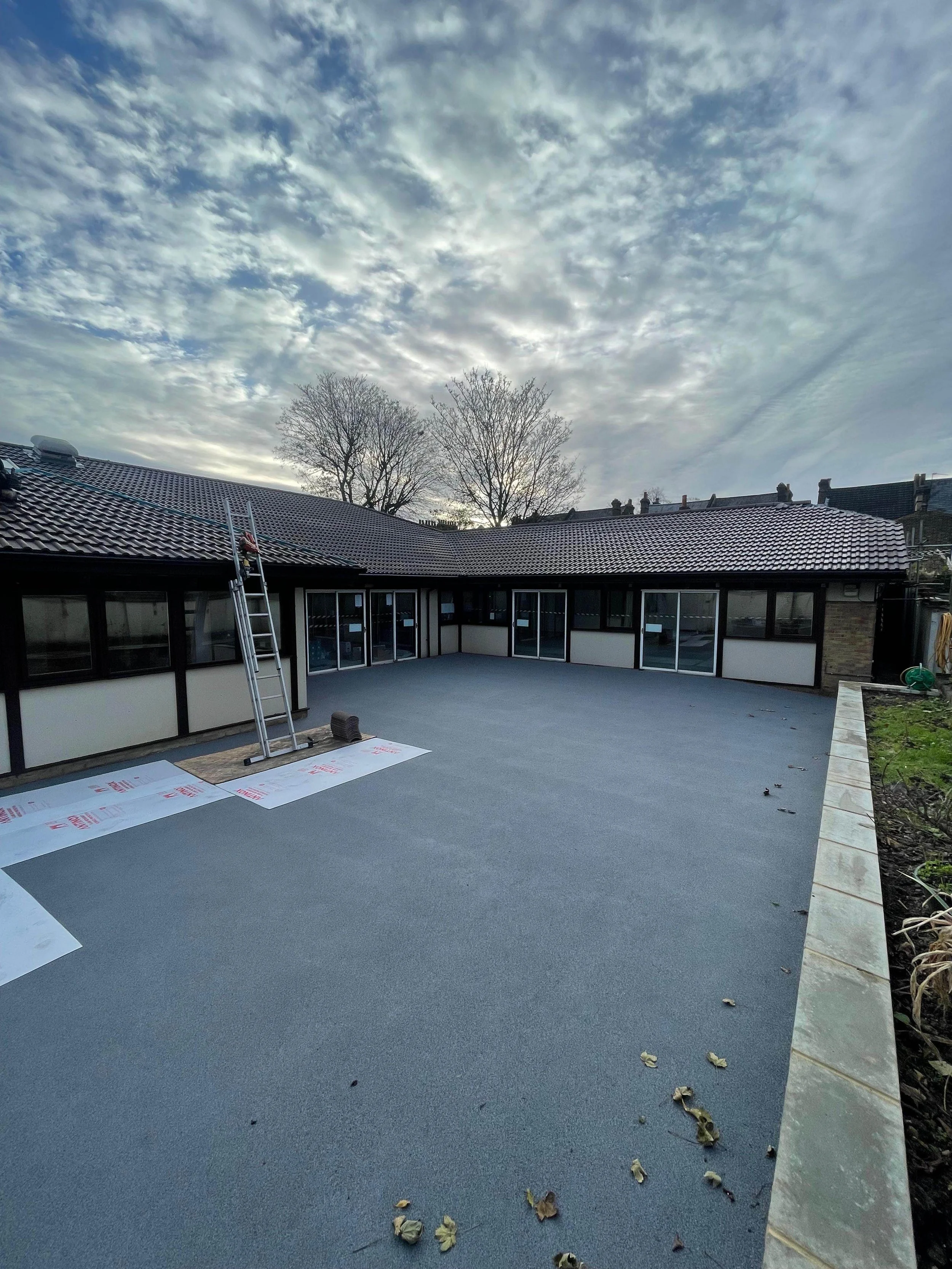 Newly completed flat outdoor patio with dark gray flooring, surrounded by a low wall and accessed through multiple glass doors, with a partly cloudy sky above and leafless trees in the background.