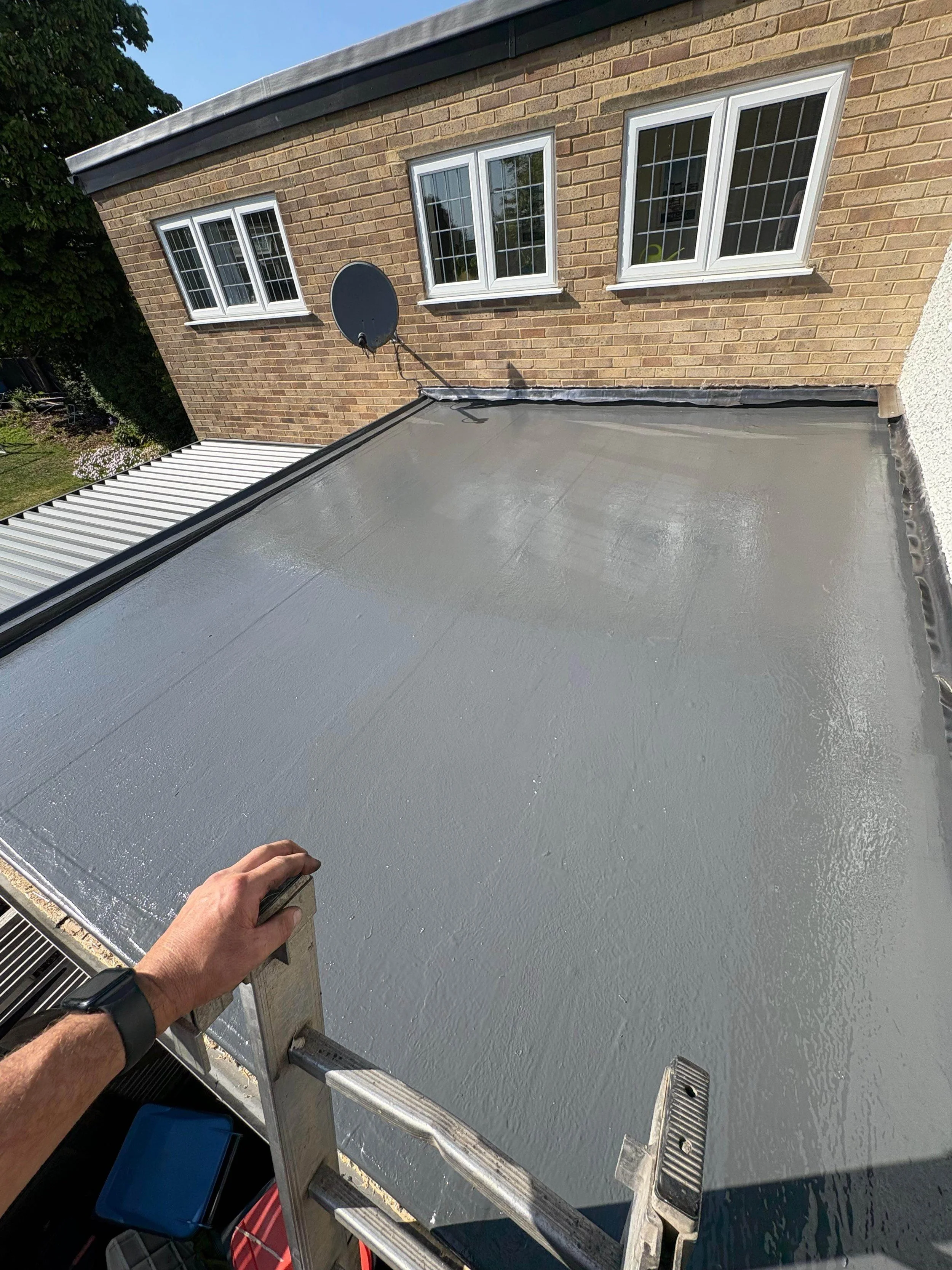 A person on a ladder applying a gray waterproof coating to a flat rooftop with a beige brick building in the background that has three windows and a satellite dish.
