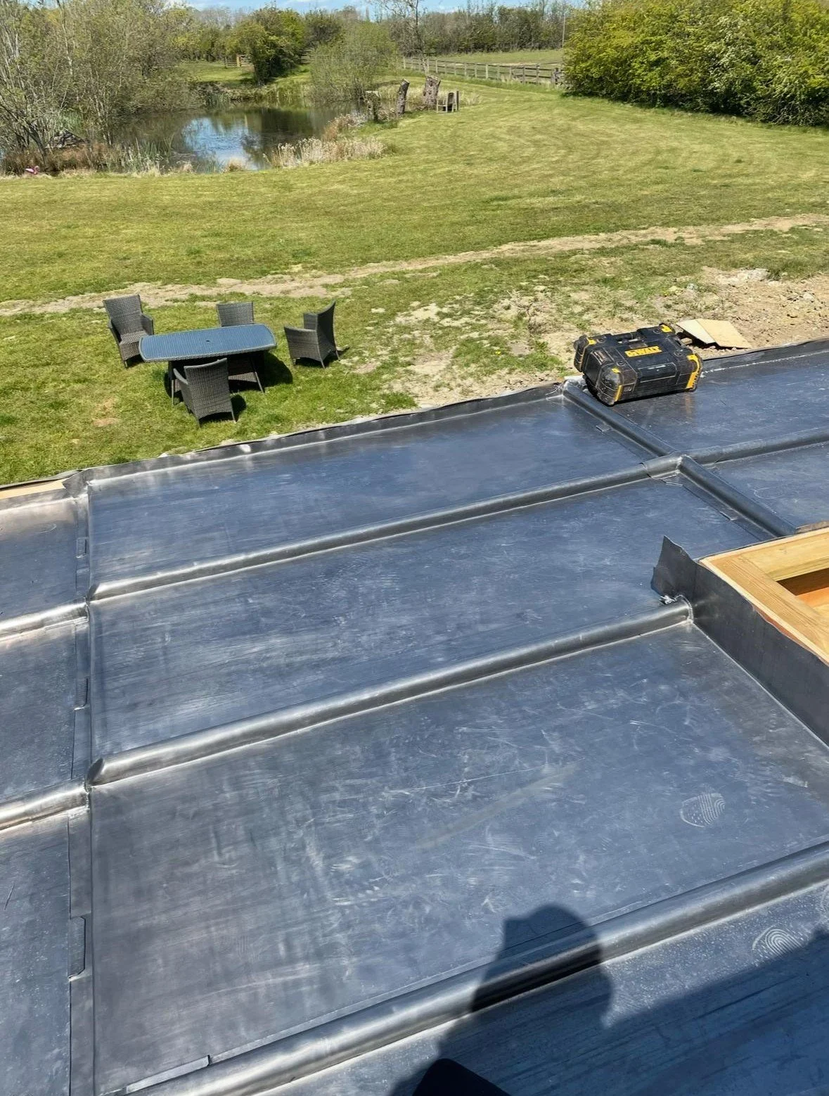 Metal roofing sheets on a building under construction, a black toolbox on the roof, and a grassy yard with outdoor furniture near a pond in the background.