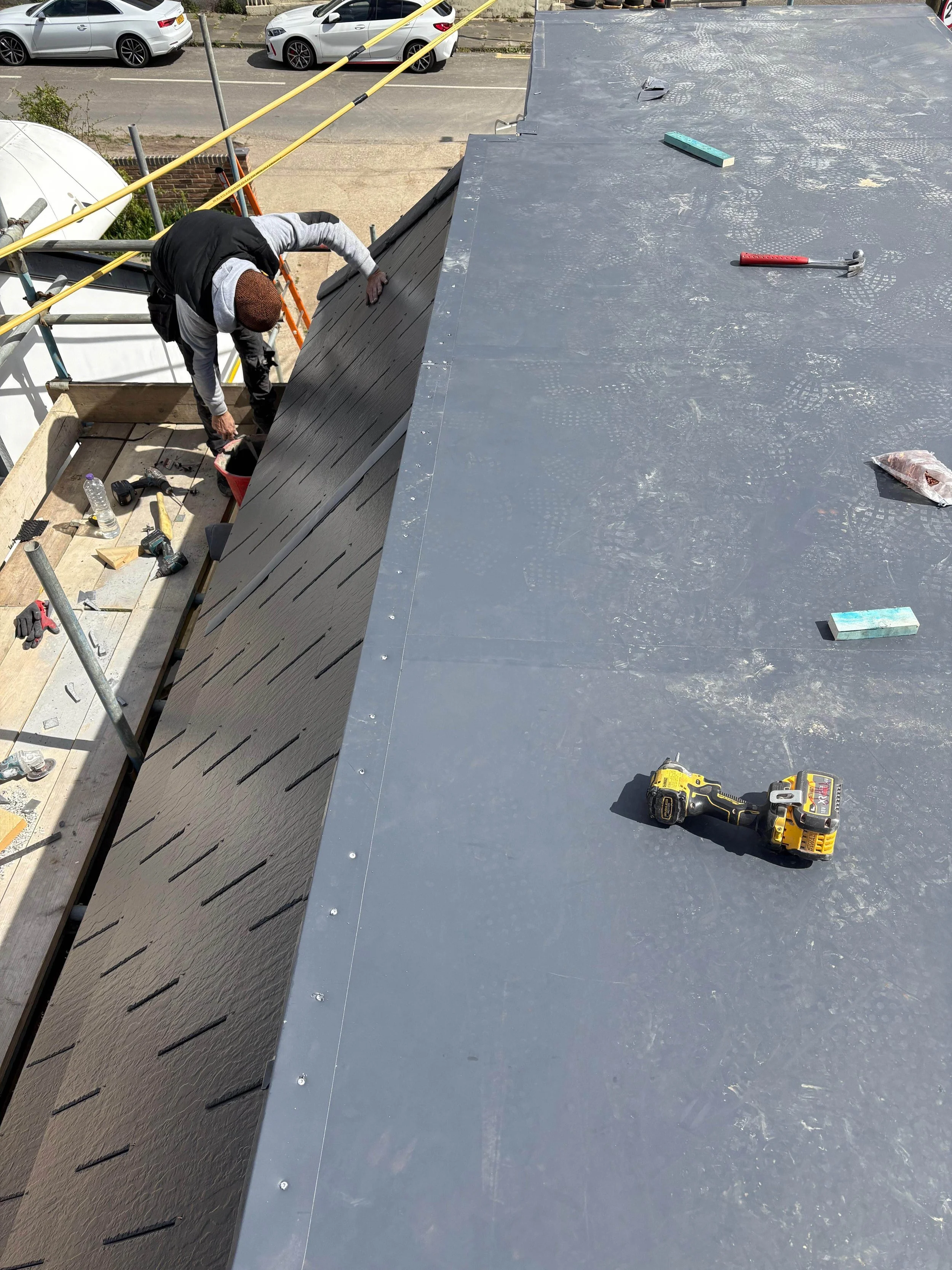 A construction worker is installing siding on a building's roof. The worker is bent over, working on the edge of the roof with tools nearby, including a cordless drill, a hammer, and a sponge. The roof is partly covered with dark material, and there 