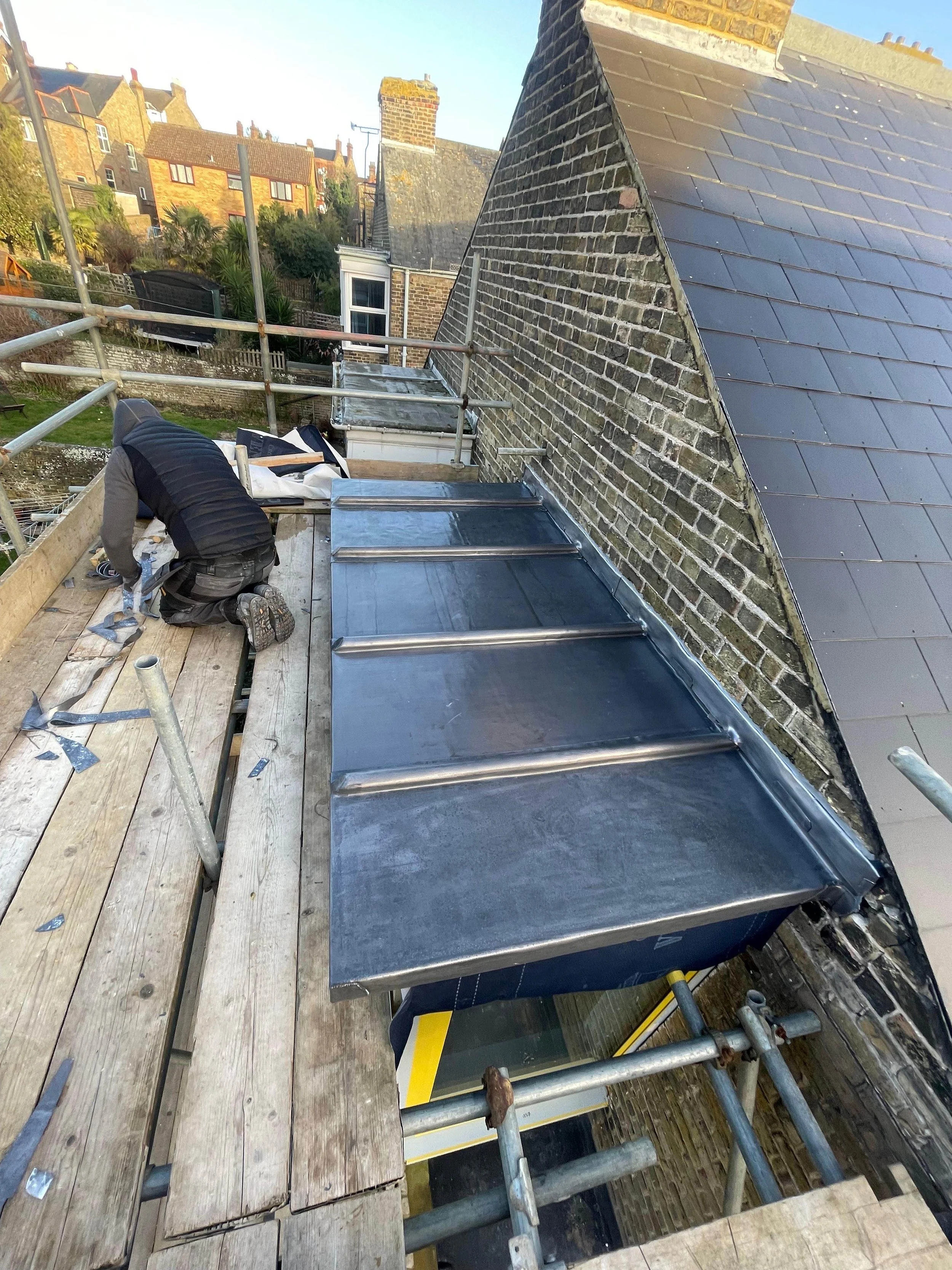 A worker installing metal roofing on a building's rooftop. The worker is kneeling, wearing a hooded jacket, surrounded by construction tools and scaffolding. The building has a brick wall and neighboring houses are visible in the background.