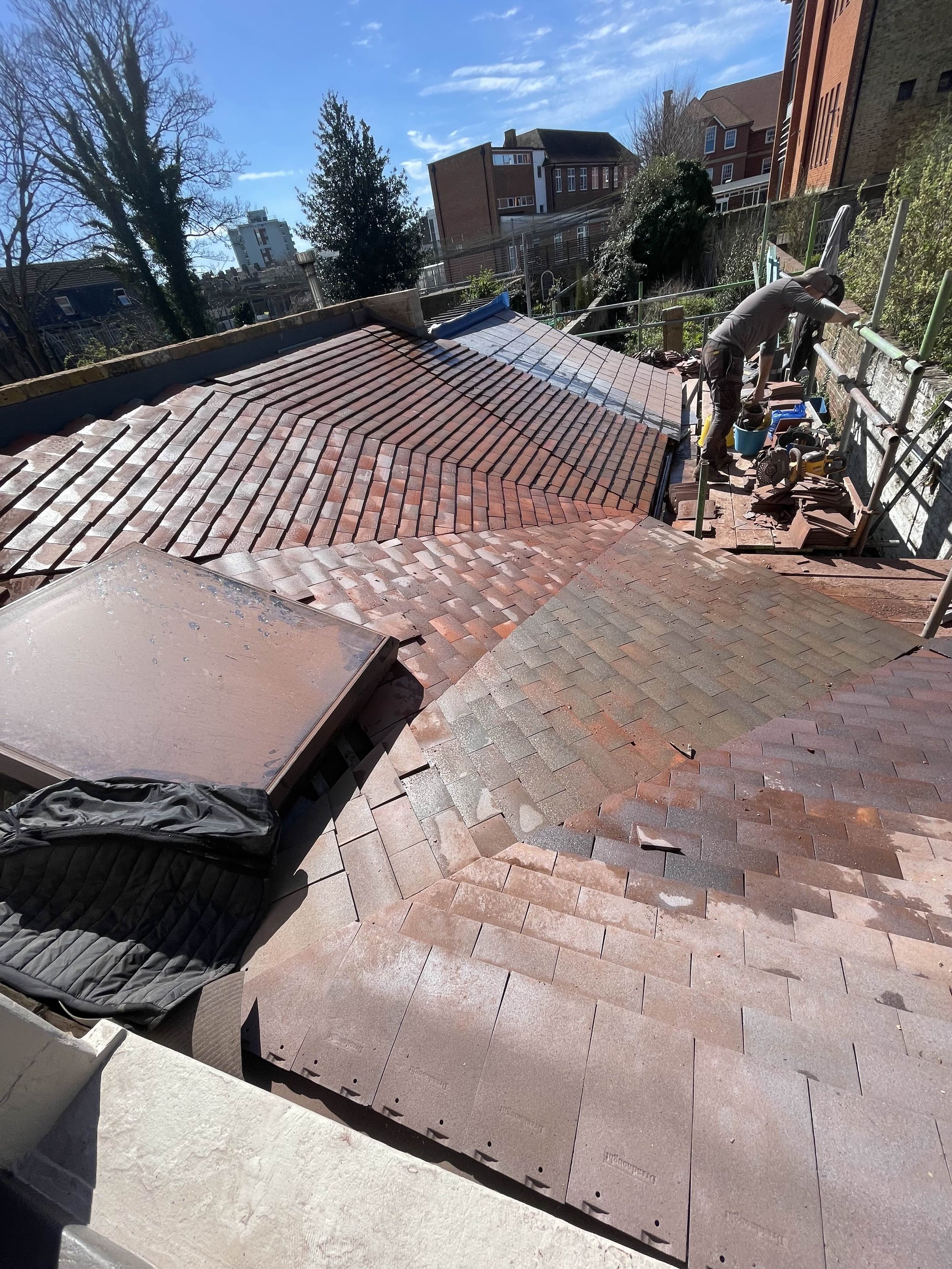 A worker on a roof installing or repairing roof tiles with various tools and materials nearby, under a clear blue sky with an urban background.