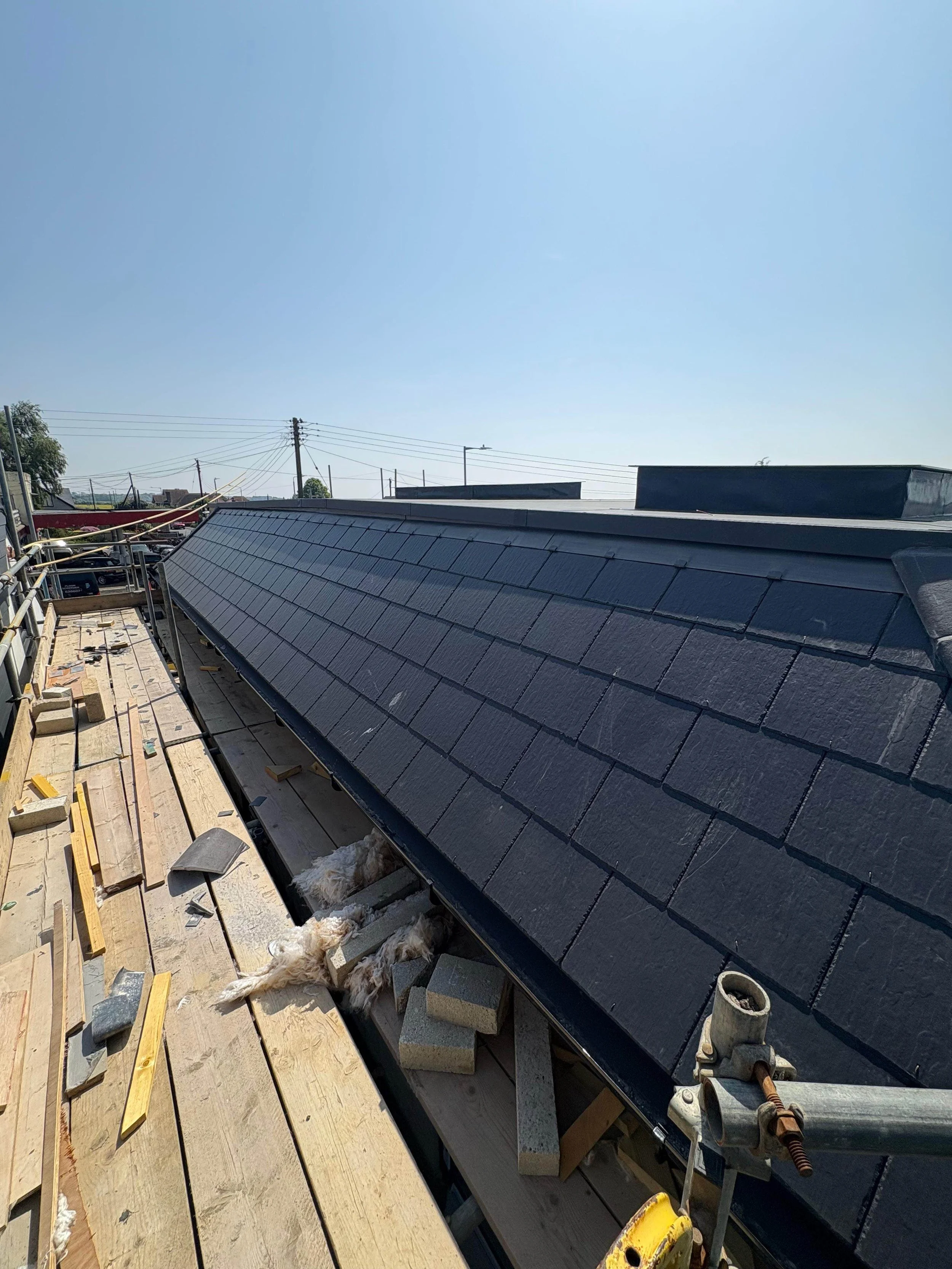 Construction site on a rooftop showing newly installed dark gray roof shingles, with construction materials, tools, and a small dog lying on the wooden platform.