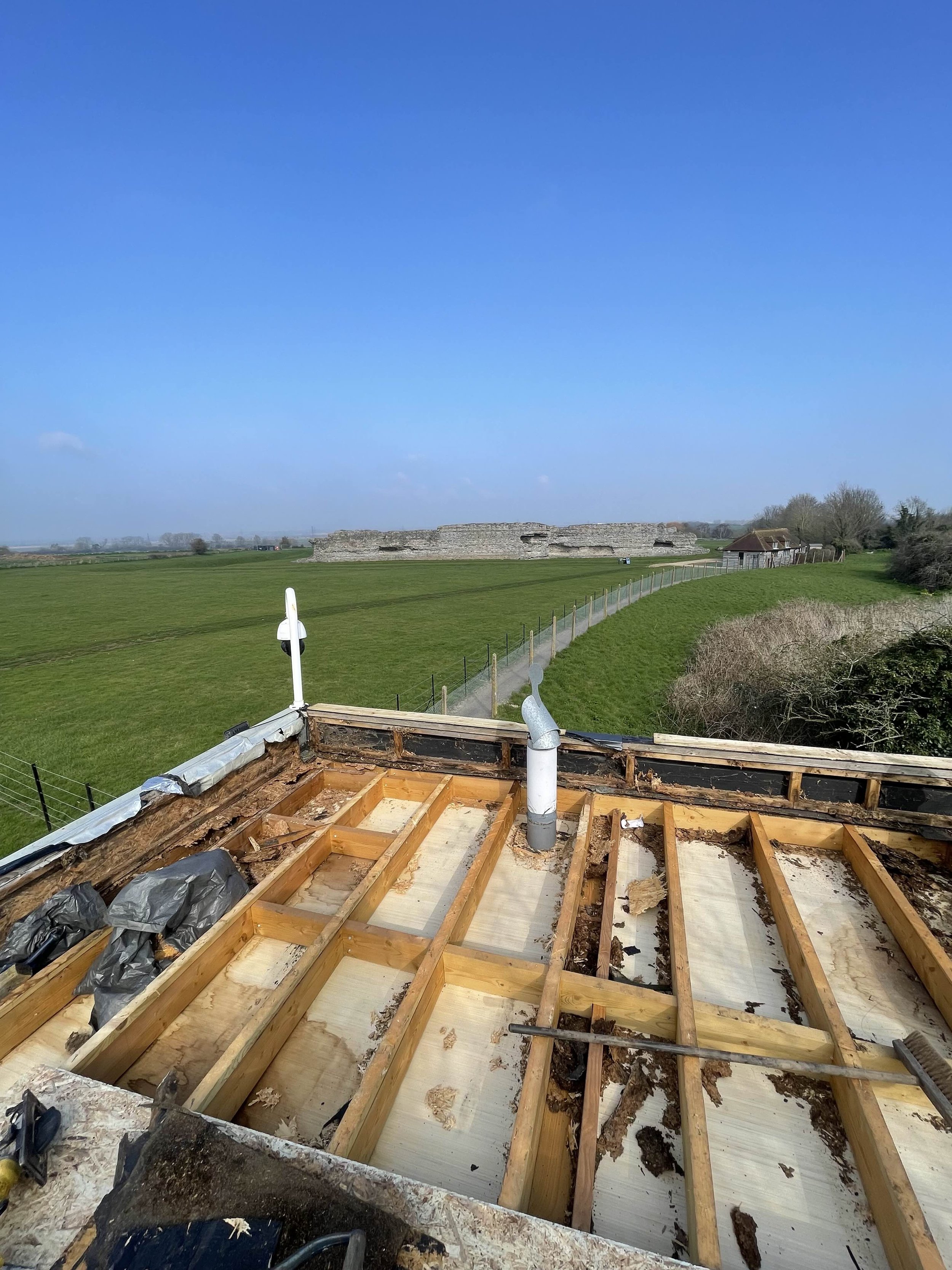 Construction site on a rooftop with wooden beams and ducts, overlooking green fields and ruins in the distance under a blue sky.
