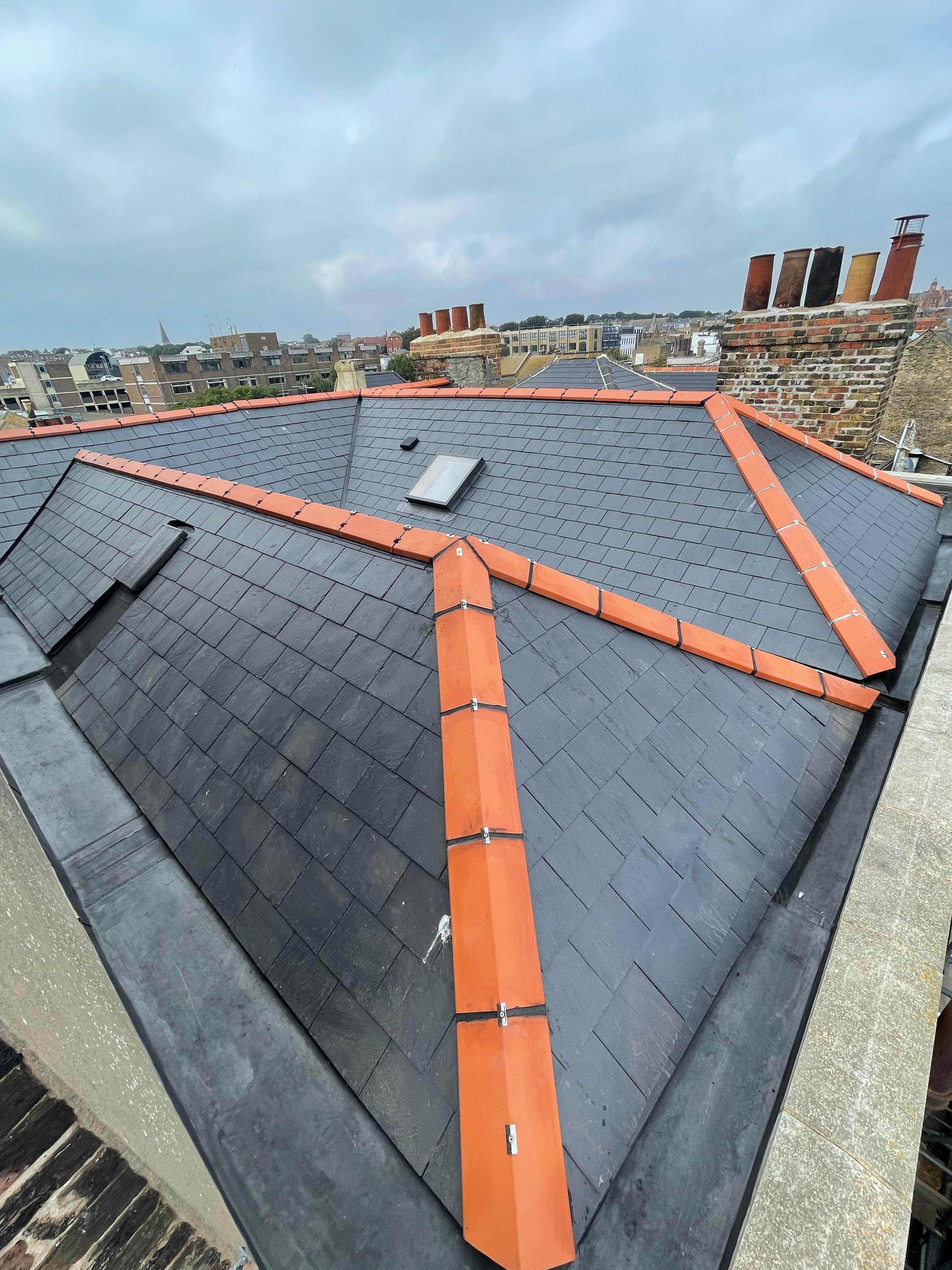 Rooftop with dark gray tiles, orange ridge tiles, and a small skylight, with a cityscape and cloudy sky in the background.