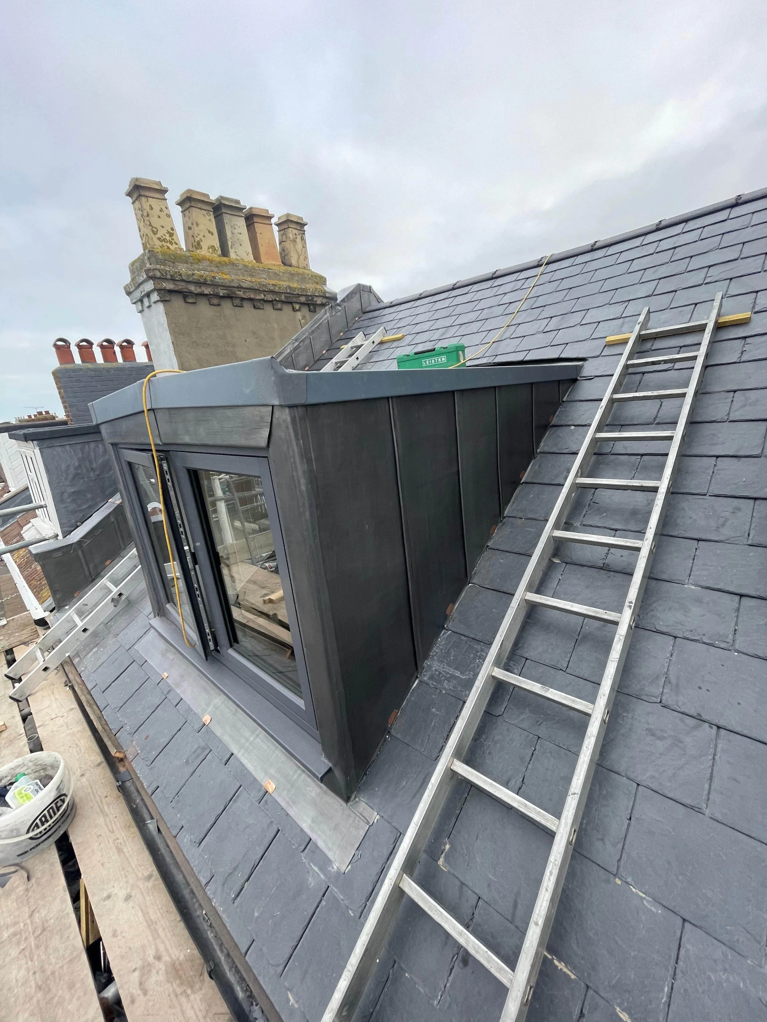 On the roof of a building, a skylight window is being installed with a ladder leaning against the roof. Tools and construction materials are nearby under an overcast sky.