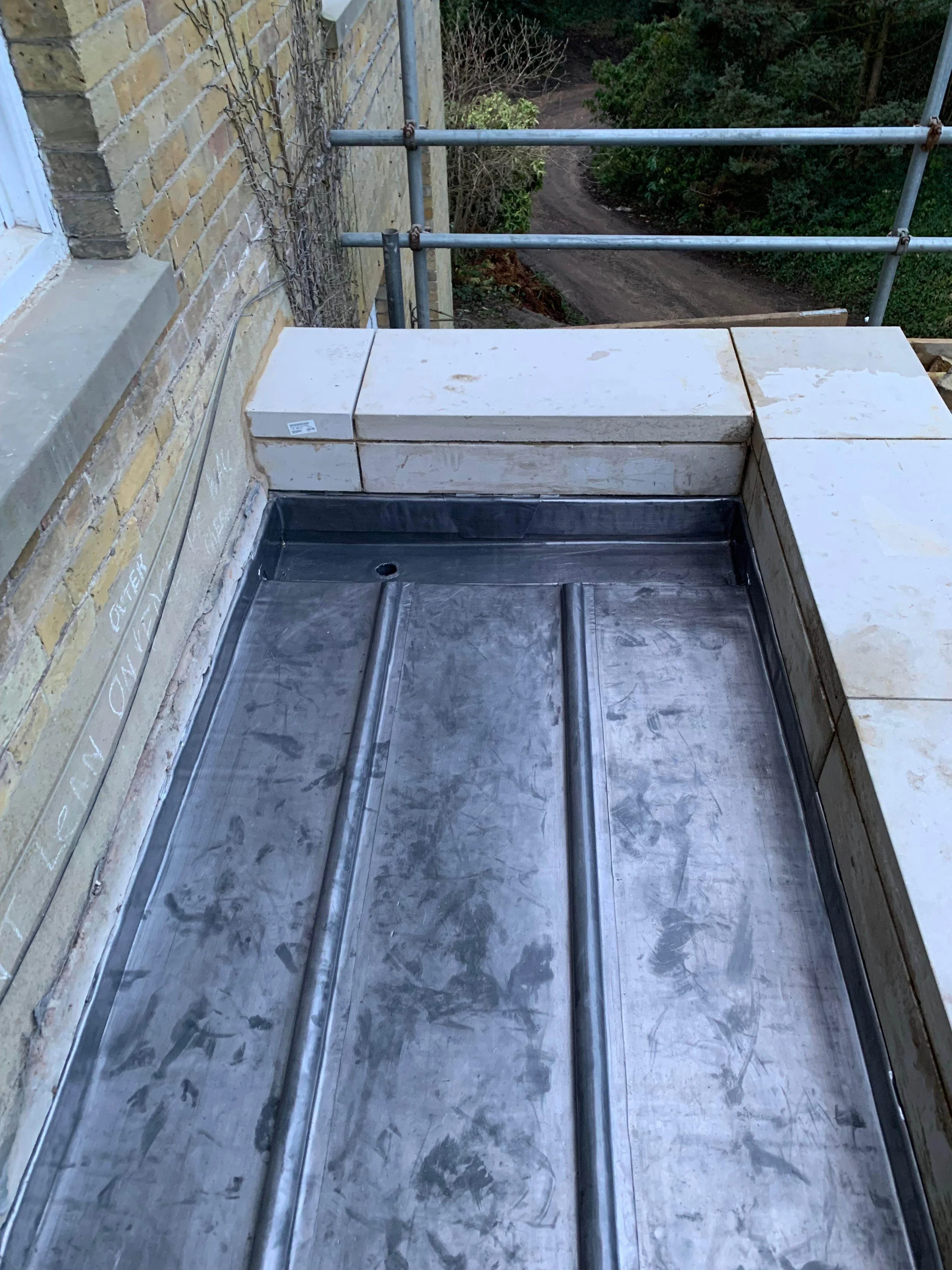 Construction site showing a metal roofing tray with white stone tiles around it, along with scaffolding and a brick wall, overlooking a dirt path and trees.